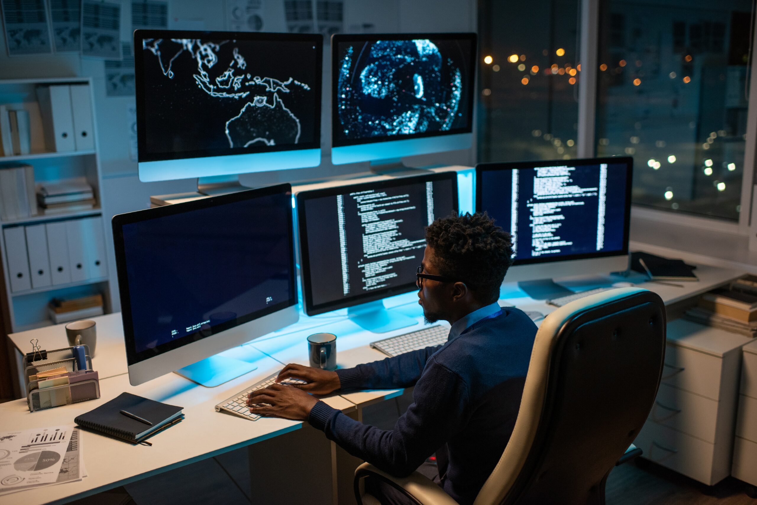 A man sitting at a desk with three monitors in front of him, providing Cybersecurity as a Service.