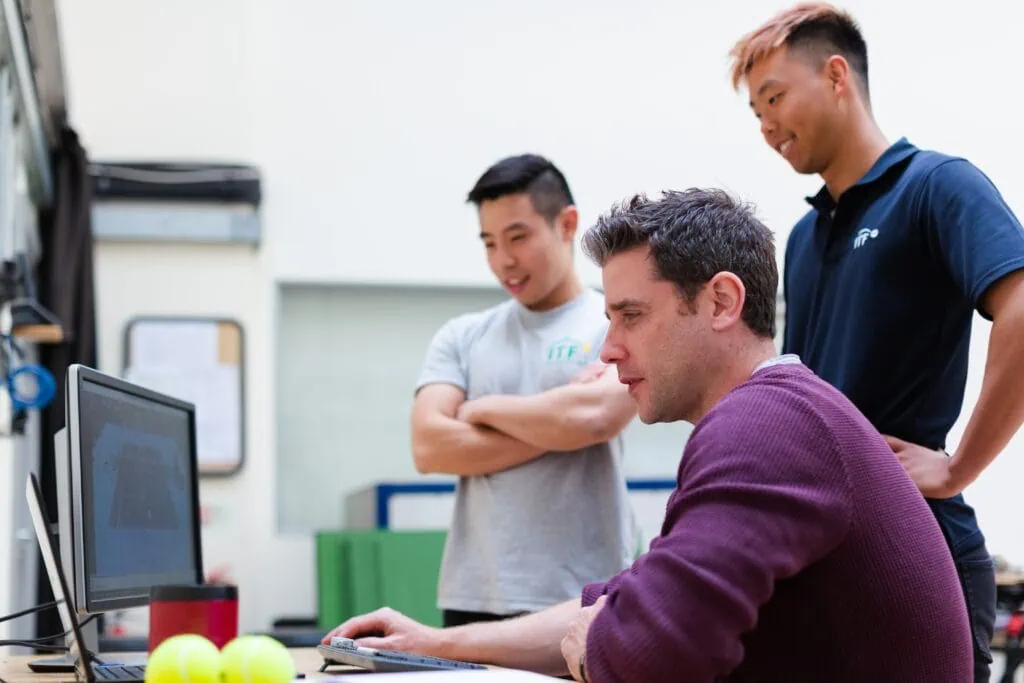Three men from a small business meticulously manage their IT services as they attentively analyze data on a computer while tennis balls sit in front of them, offering a brief moment of recreation.