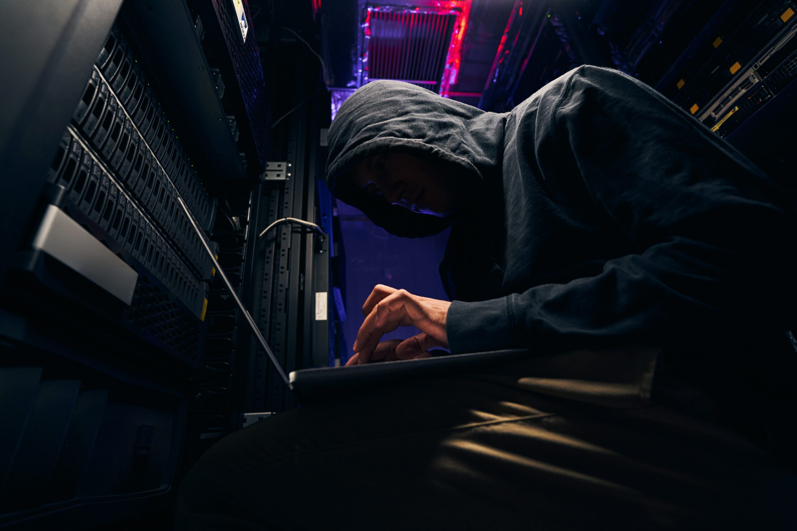A man in a hoodie using a laptop in a server room, sending a phishing email.