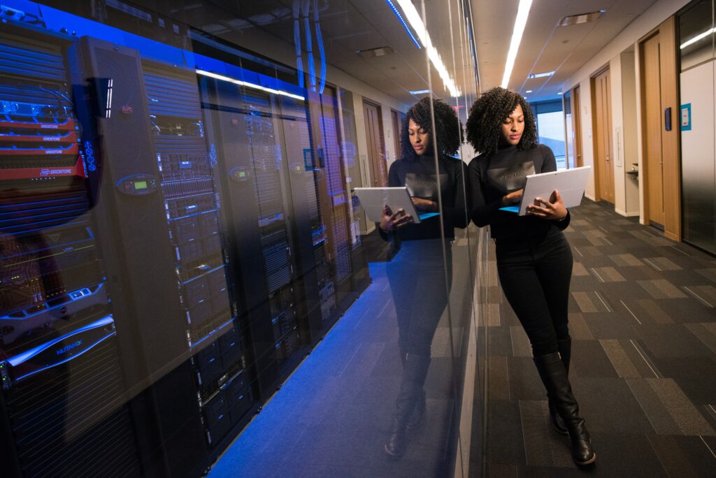 A woman is standing outside a server room providing IT consulting and services, while looking at a laptop.