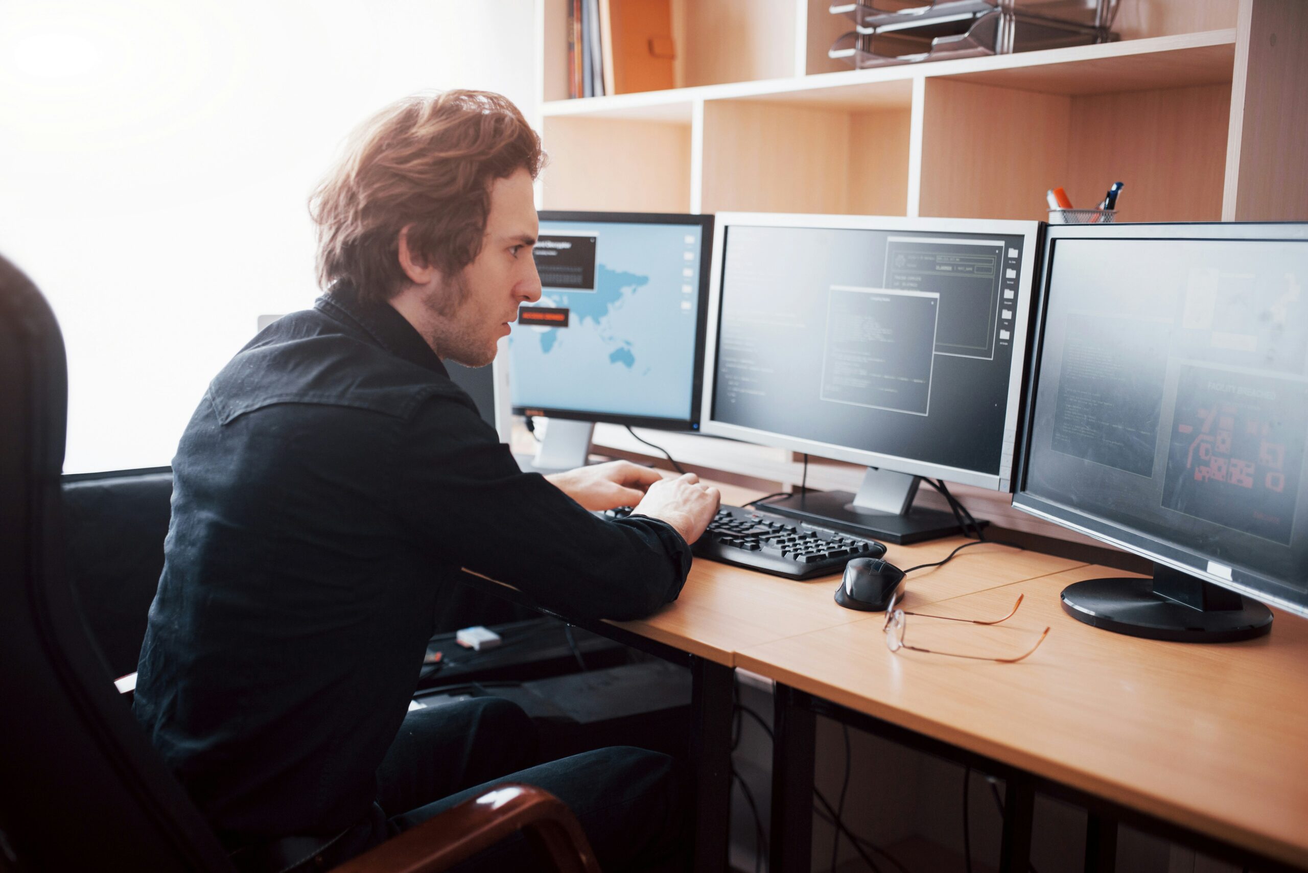 A man sitting at a desk, diligently monitoring multiple computer screens to survive potential cyber attacks.