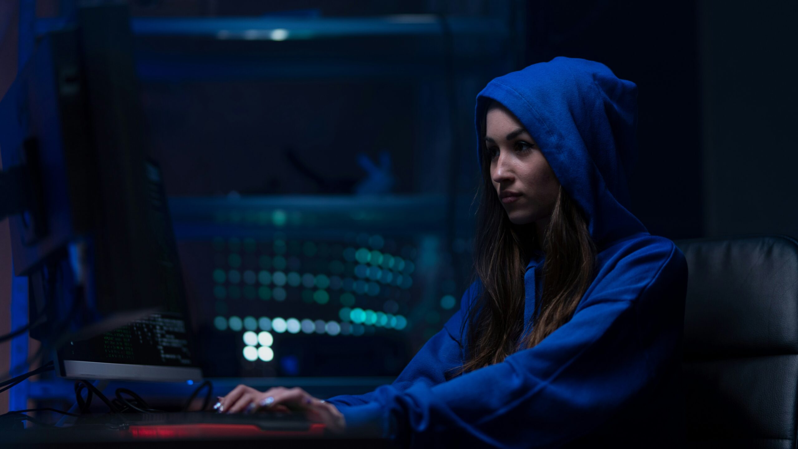 A girl in a blue hoodie sitting at a computer exploring vulnerabilities in computer systems.