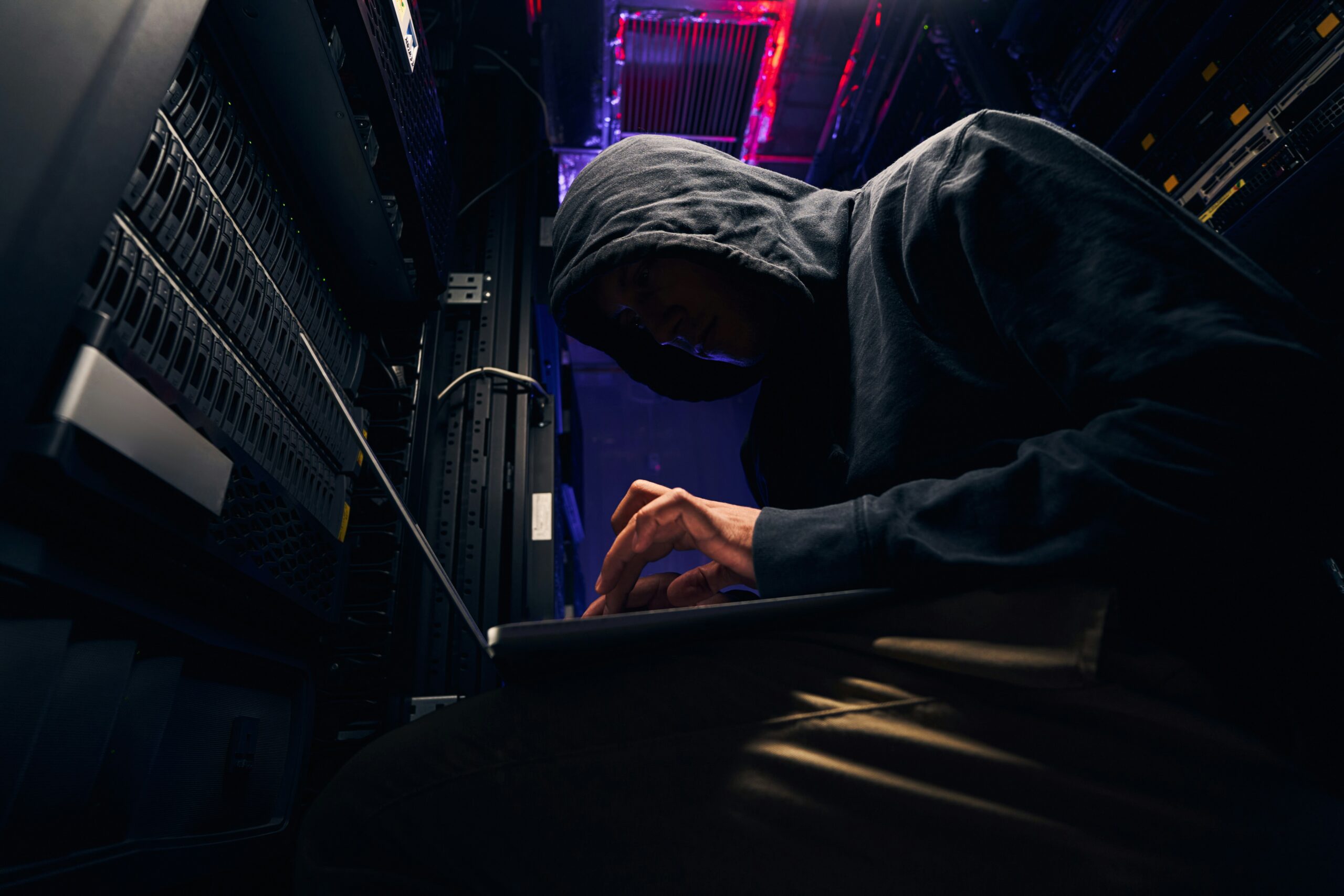 A man in a hoodie using a laptop in a server room, presumably hacking someone who does not know how to protect themselves from hackers.