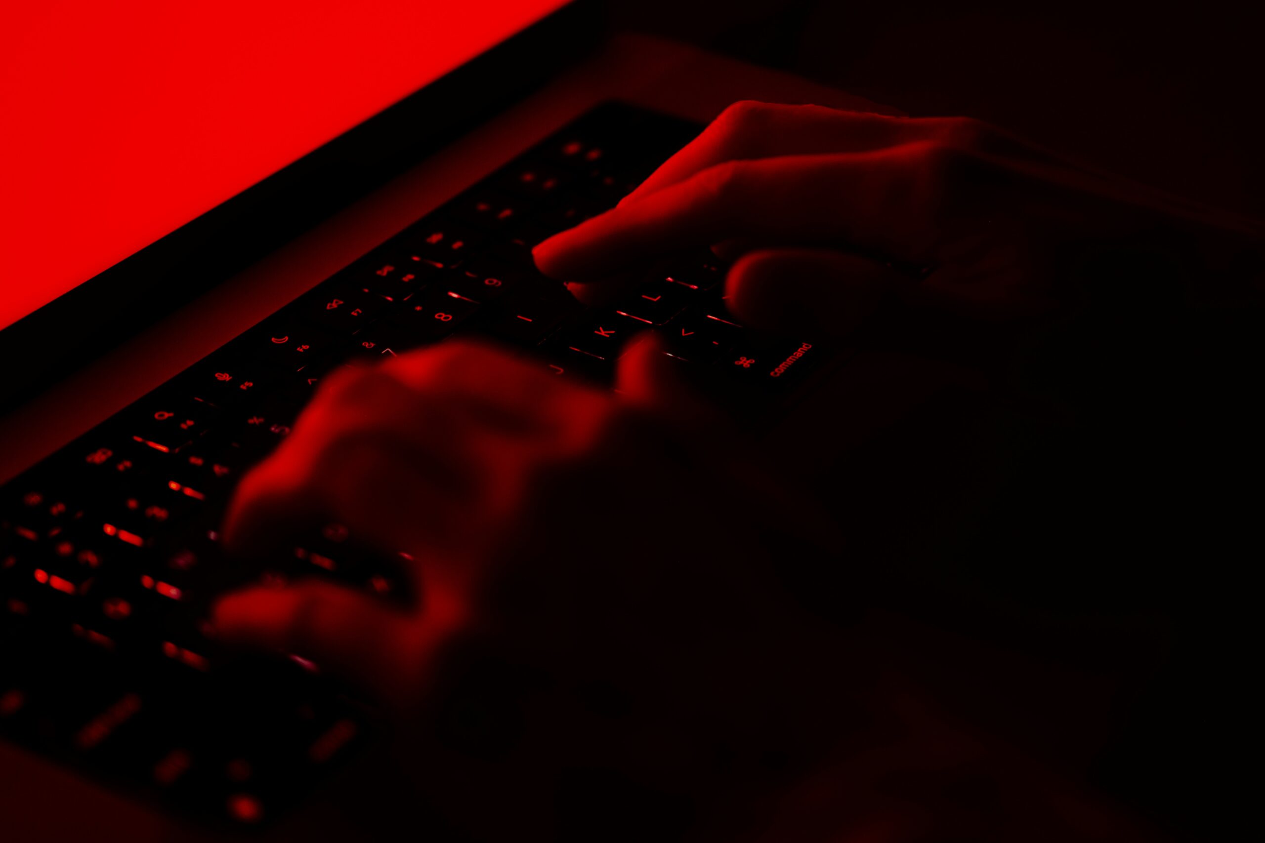 A person typing on a laptop with a red light behind them, researching how to protect themselves from hackers.