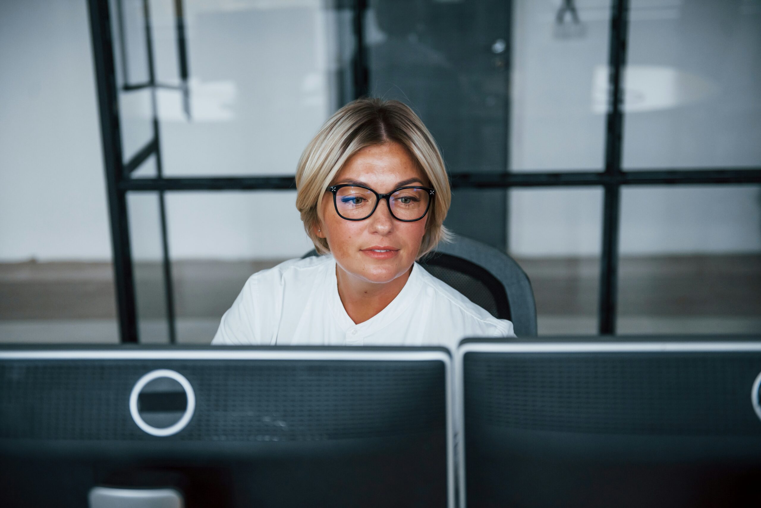 A focused woman with glasses sitting at a desk with dual monitor screens in a modern managed IT services office setting.