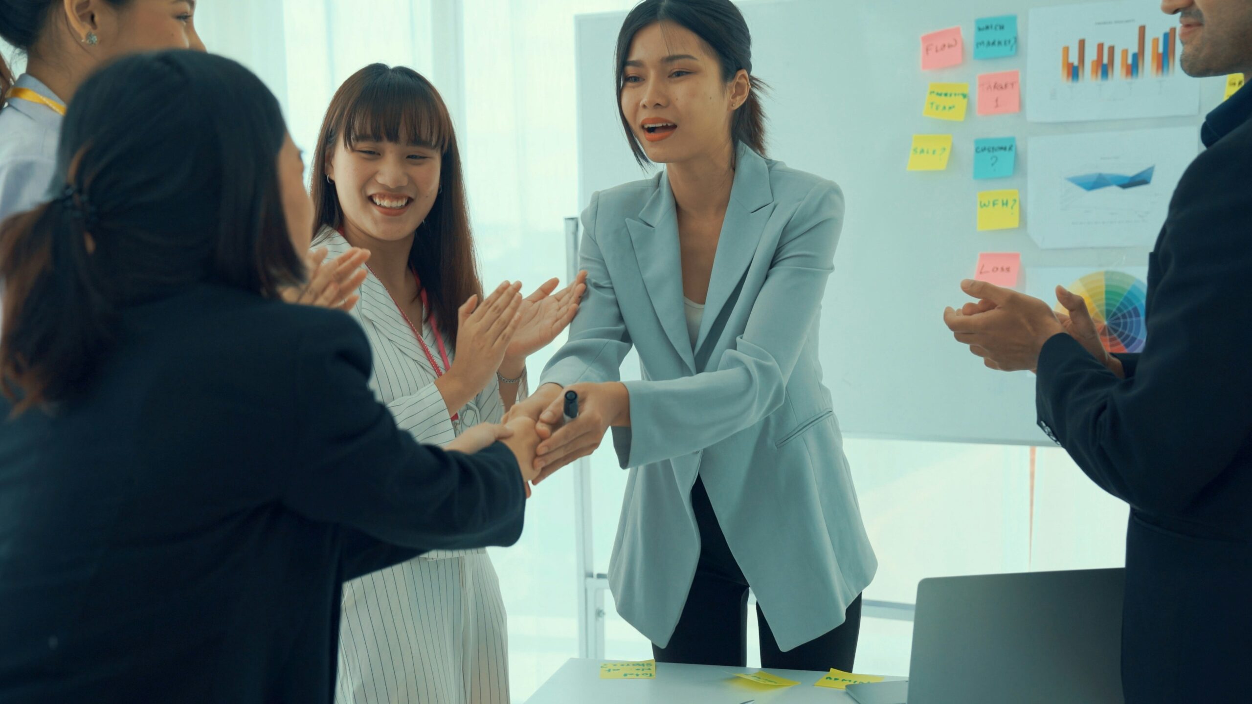 A group of five diverse business professionals celebrating the successful implementation of IT service management principles and practices in an office, clapping and shaking hands near a whiteboard with charts.