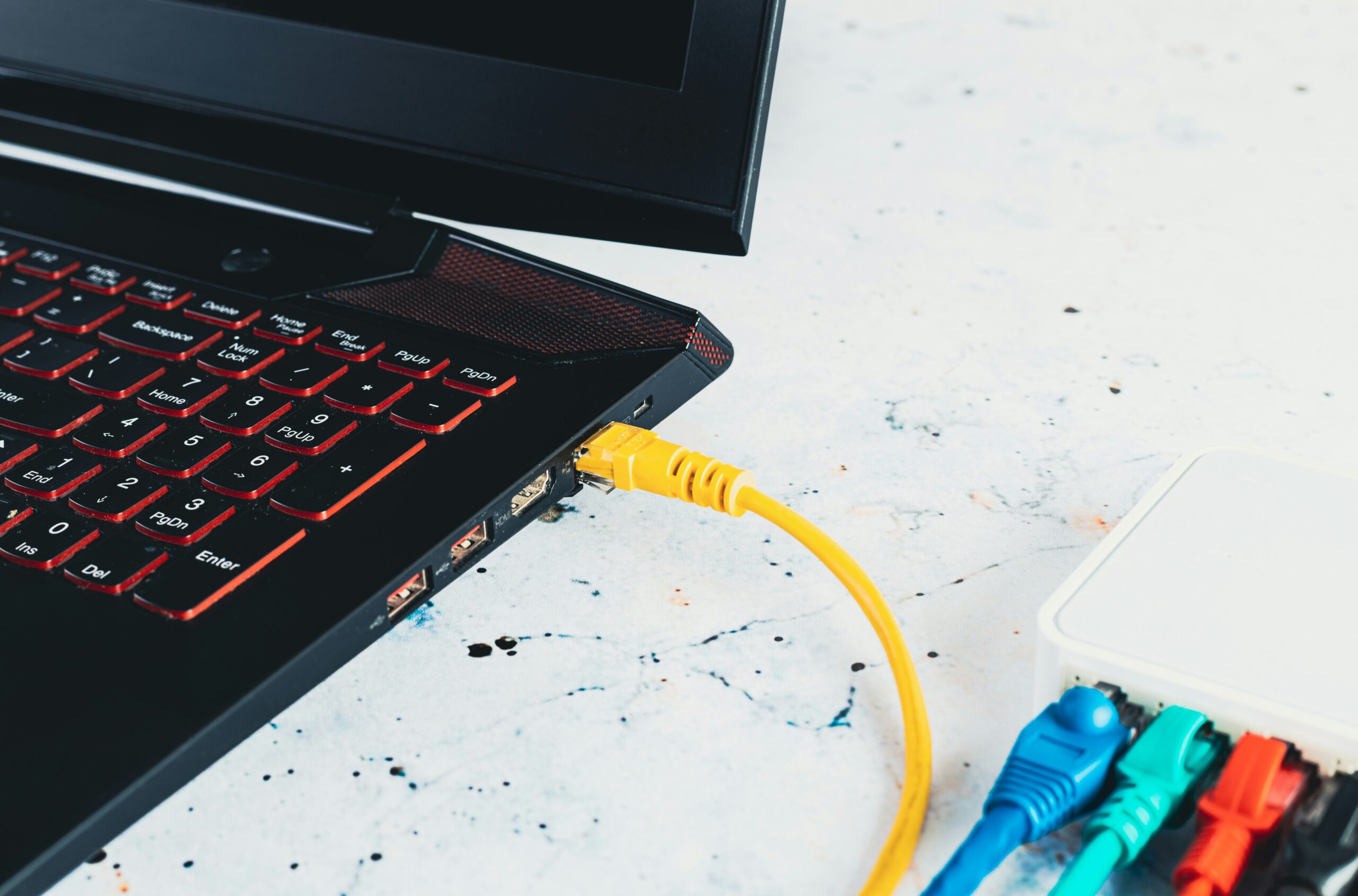 A laptop with a glowing red keyboard connected to a yellow ethernet cable, provided by an IT service management provider, next to a box of assorted colored network cables.