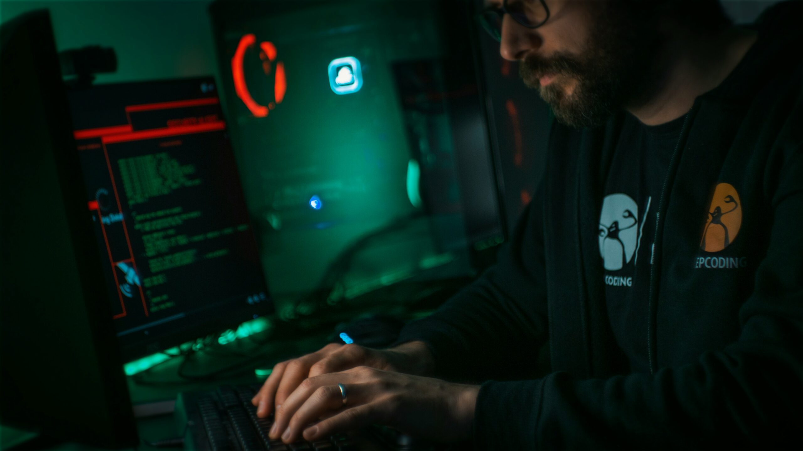A man with a beard coding on a computer in a dimly lit room with green lighting, providing MSP cyber security services.