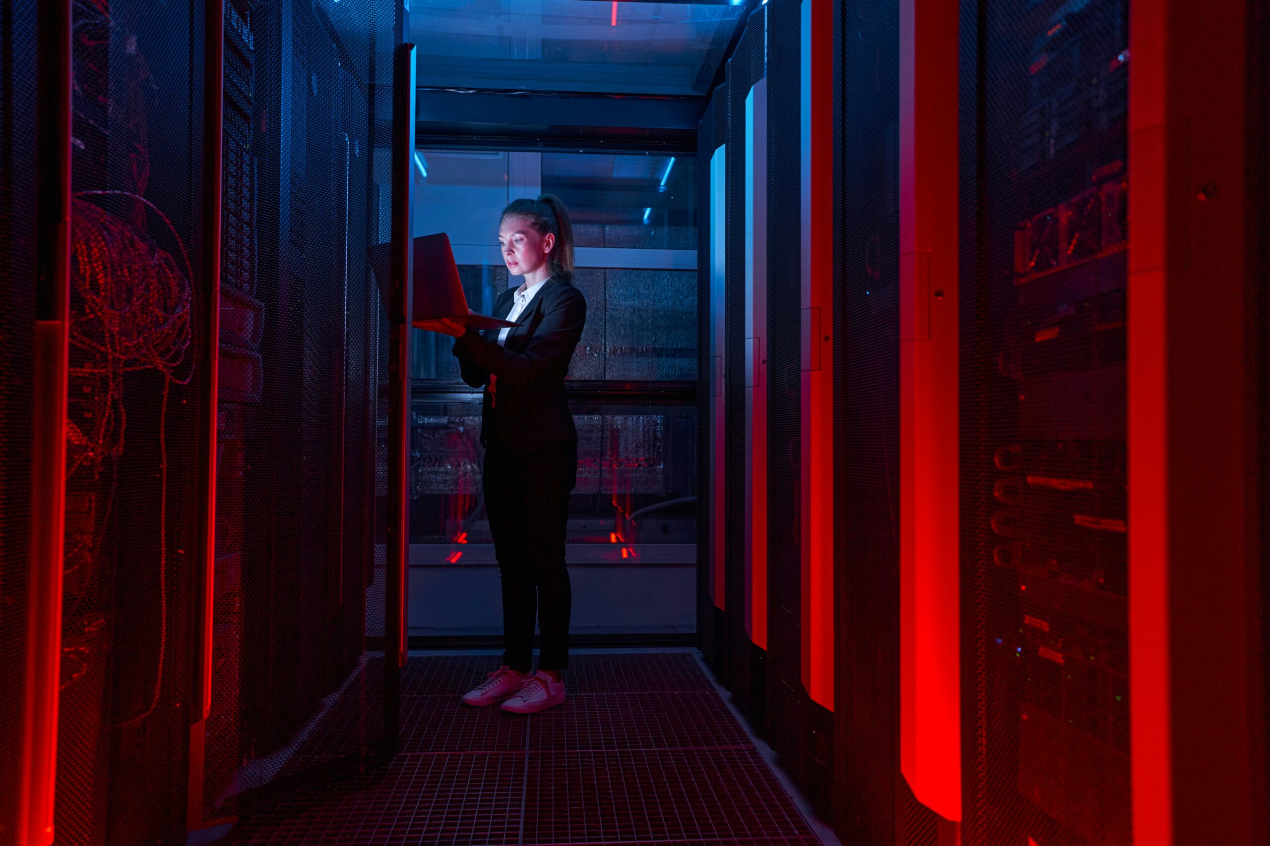 A person holding a laptop stands between server racks illuminated by red lights in an IT consulting service data center.