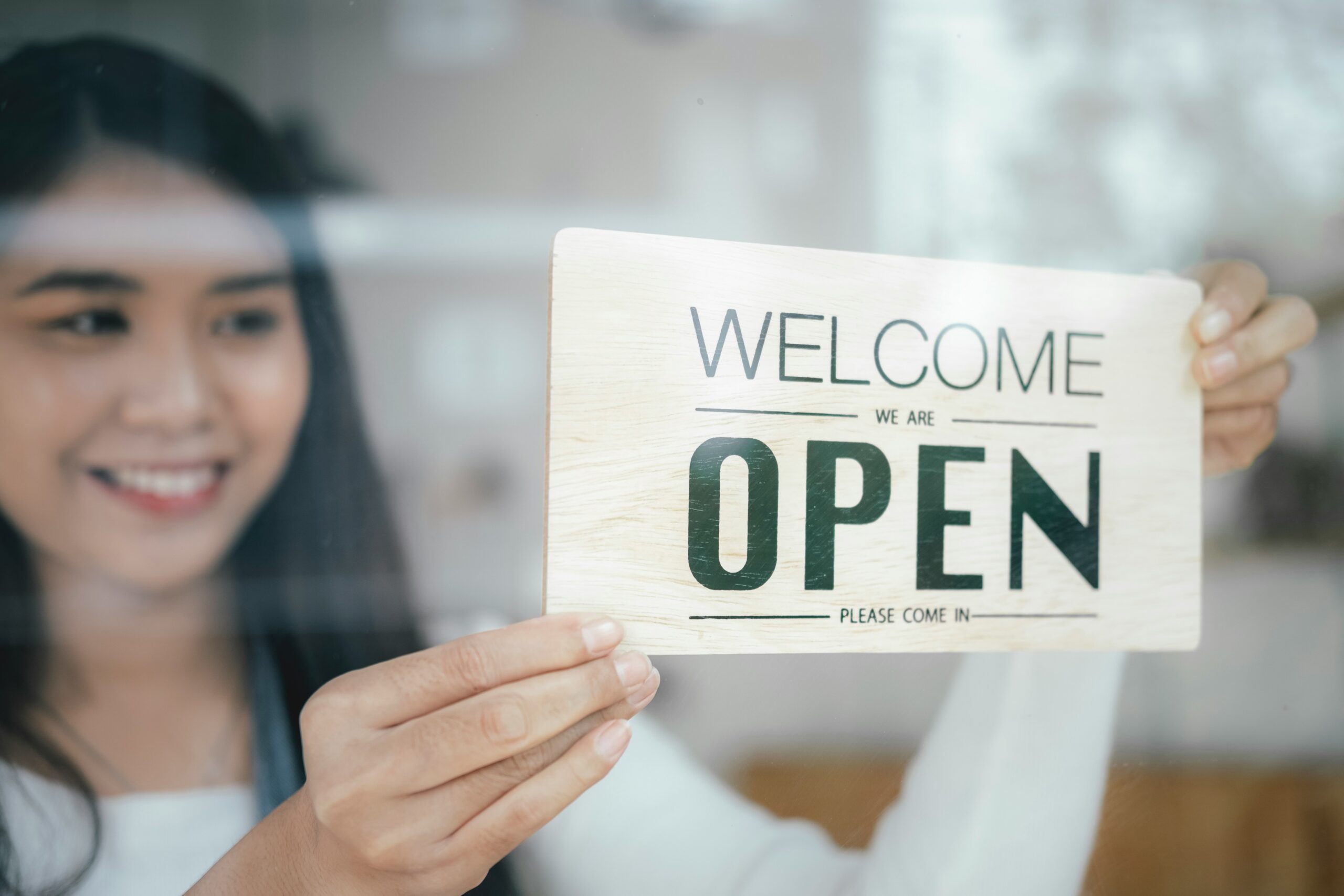 A person holding a "welcome we are open" sign on a glass pane, offering managed IT services for small businesses.