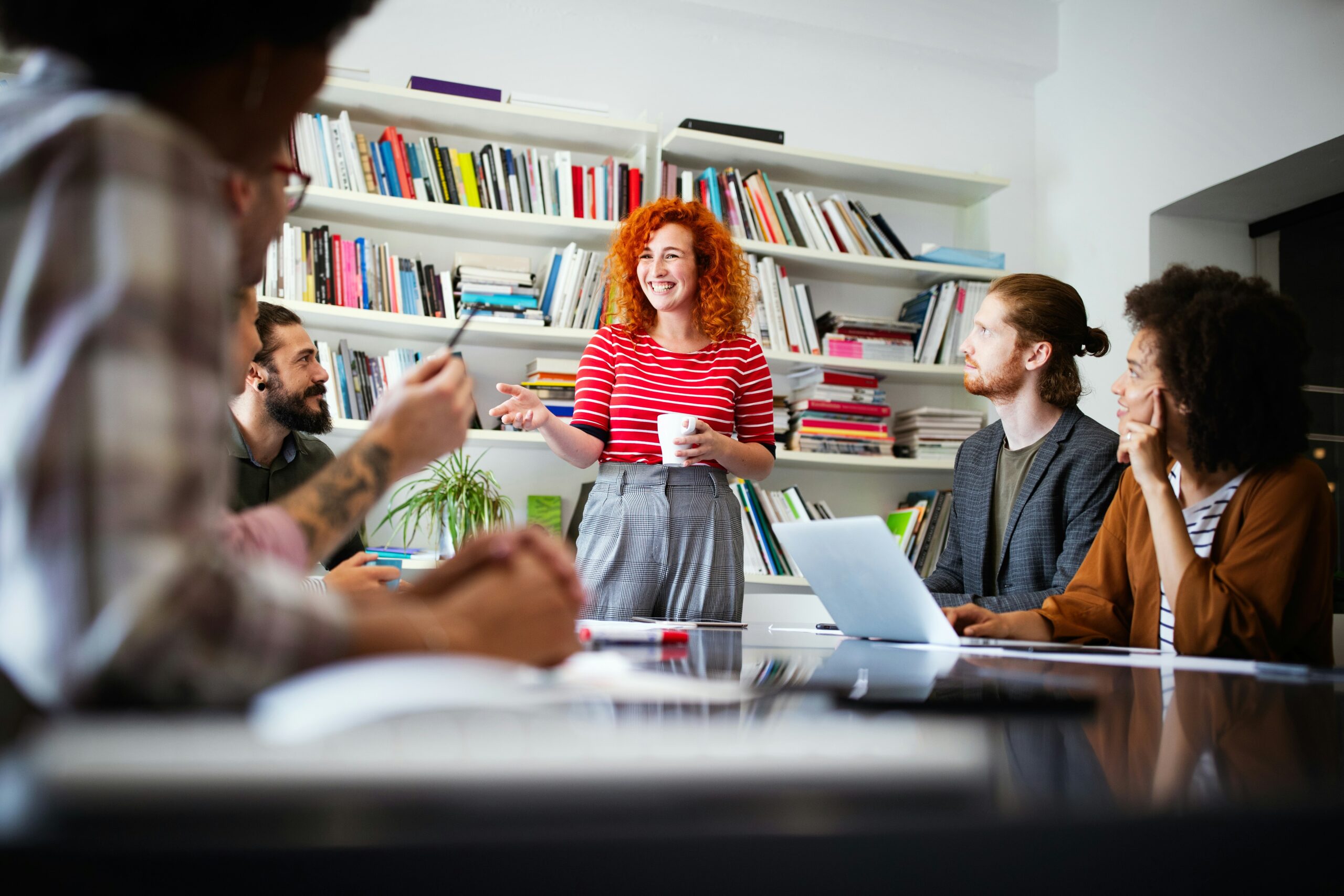 A red-haired woman in a striped shirt presents on what does an IT strategy consultant do at a meeting with four colleagues in a library.