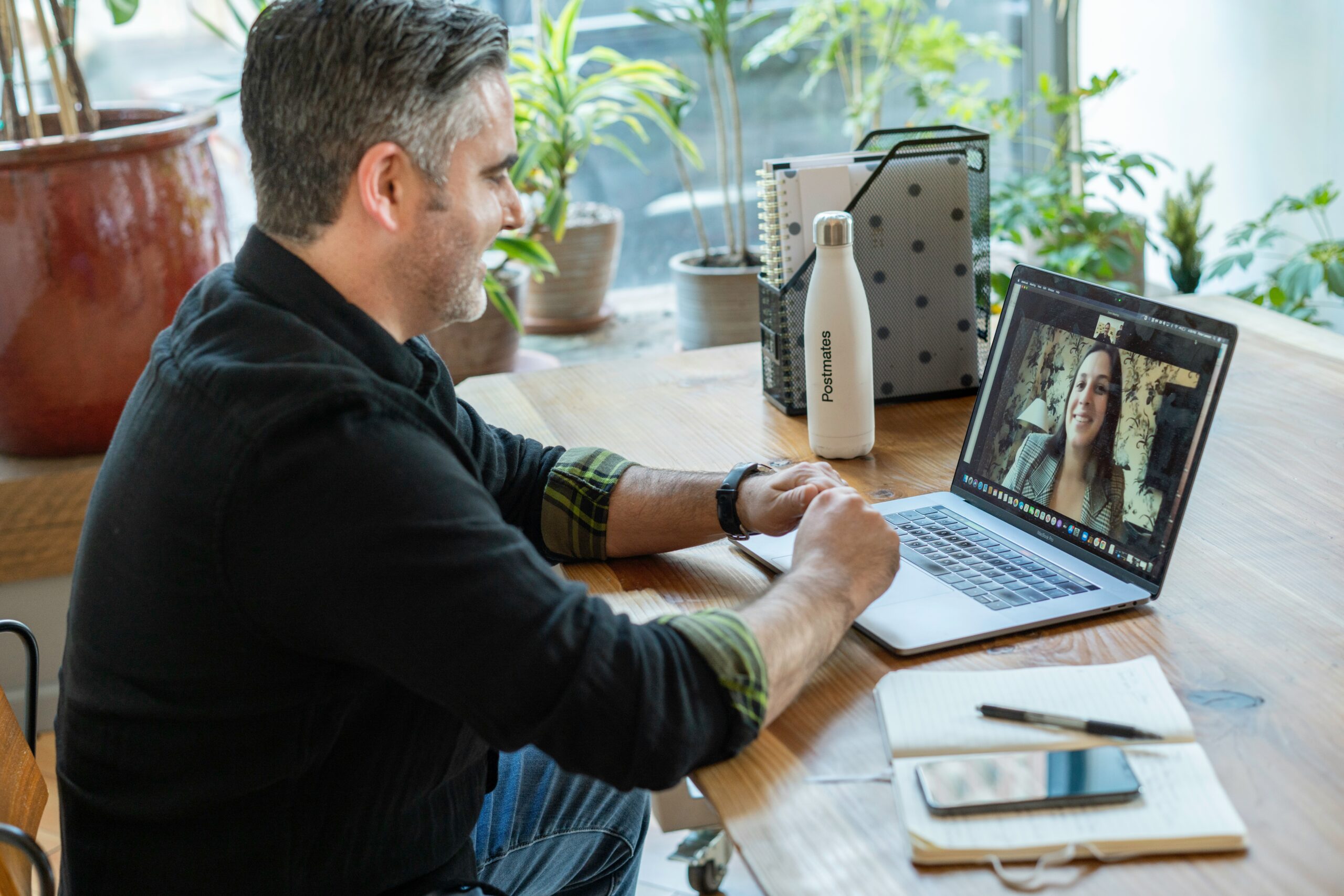 A tech consultant in a casual shirt sits at a wooden table, having a video call on his laptop with a client, surrounded by plants and notebooks.