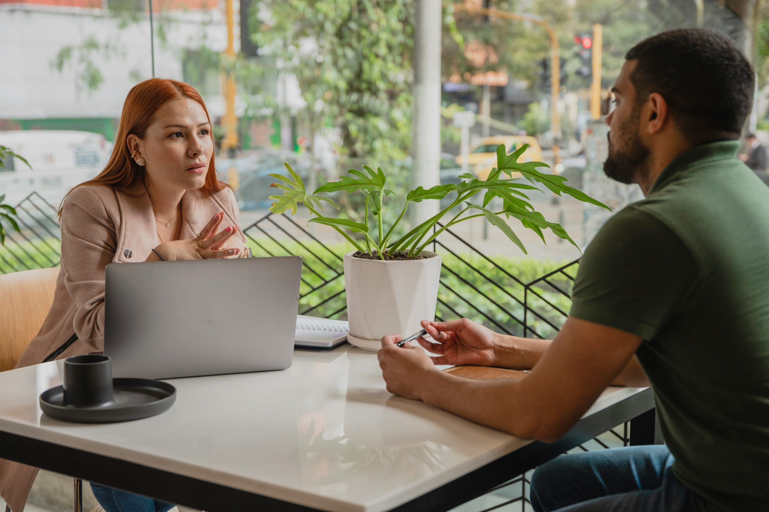 A woman and a man sit at a table with a laptop and a plant between them in a cafe, engaged in a serious conversation about what makes a good IT consultant.