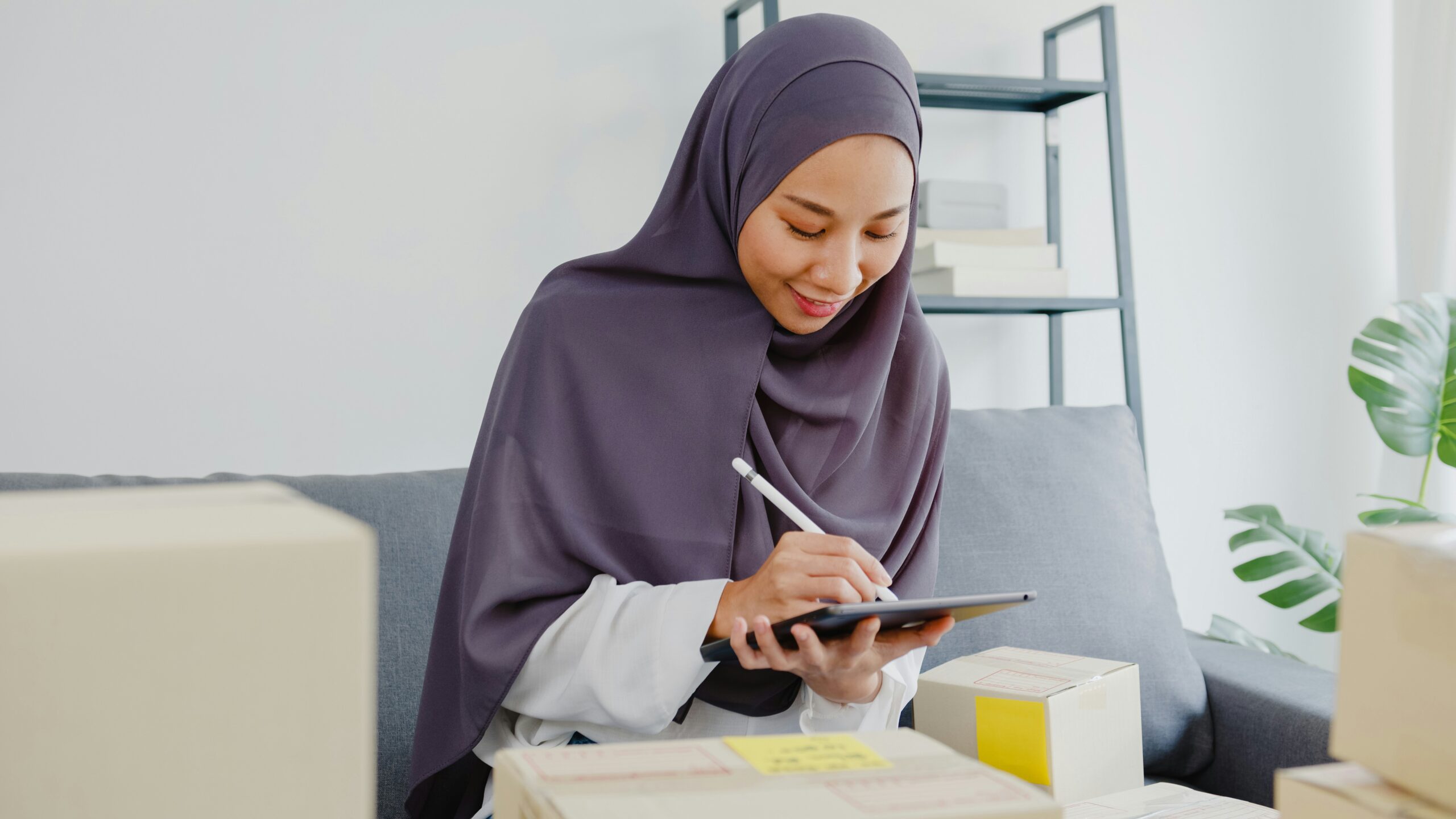 A woman in a hijab using a digital tablet and stylus while sitting on a sofa surrounded by packages and office supplies, researching what does an IT strategy consultant do.