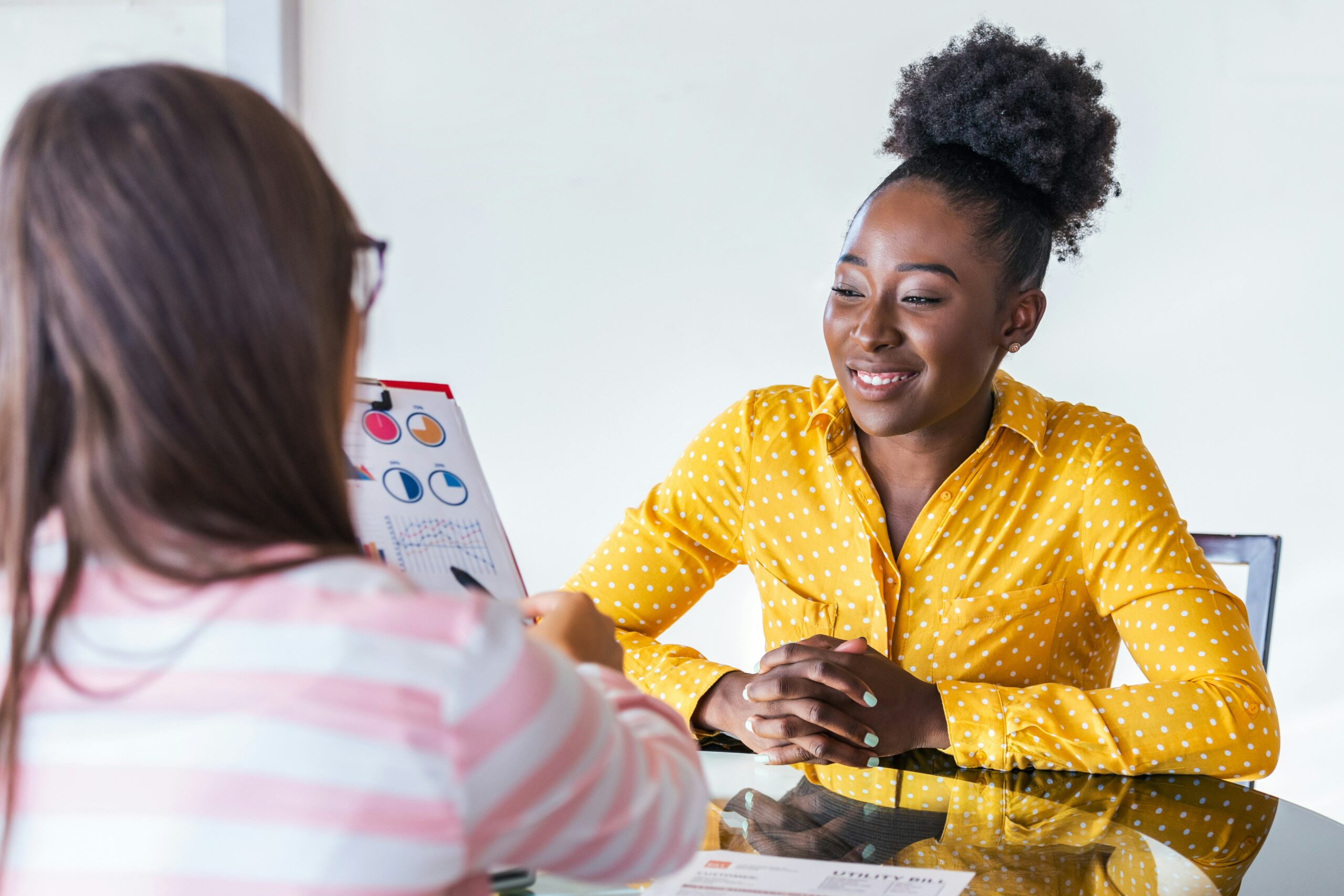 A woman in a yellow polka-dot shirt smiles while talking to a girl holding an educational book at a table, explaining what is a technical consultant.