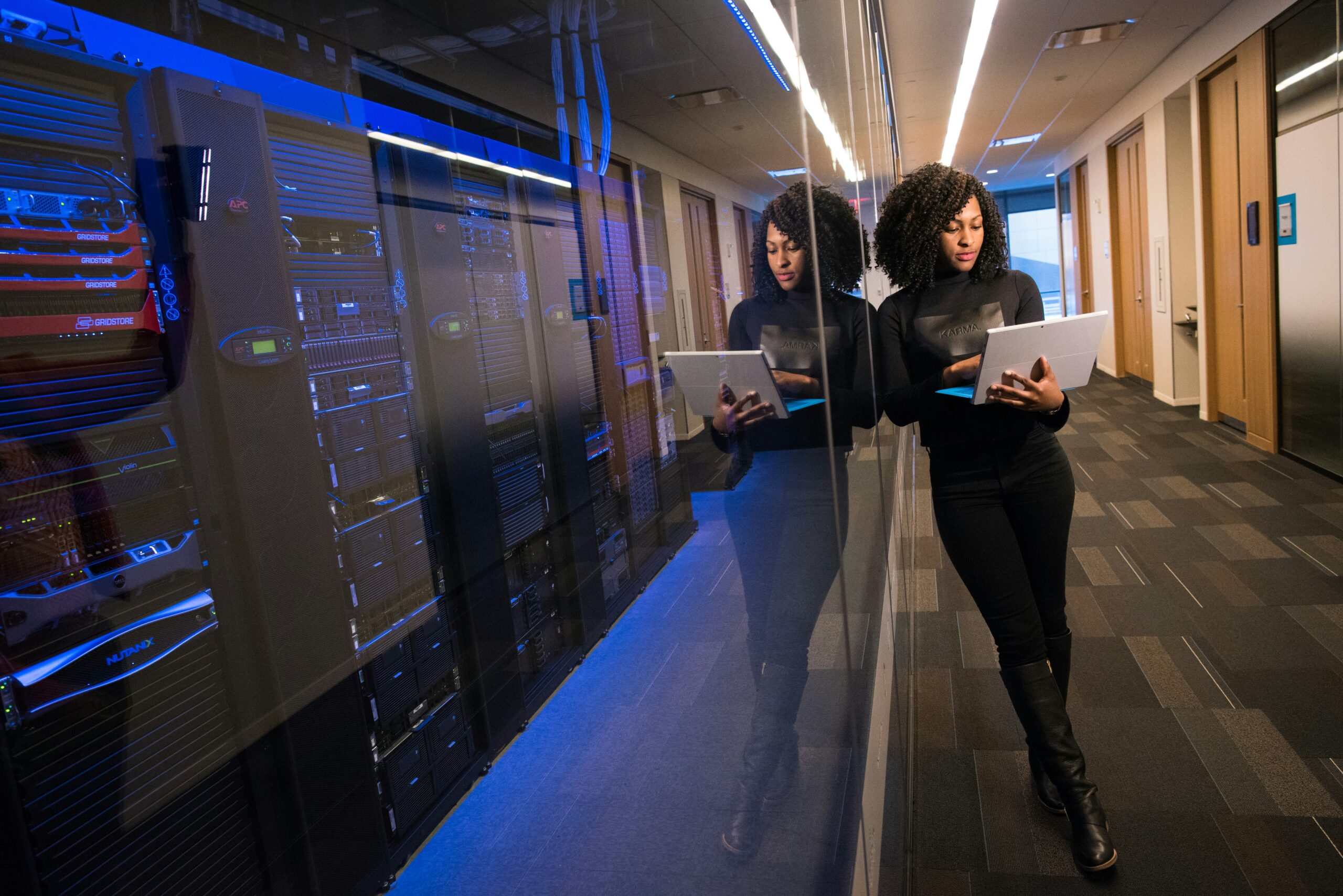 A woman using a tablet while walking through a service provider's server room with illuminated server racks on the left.