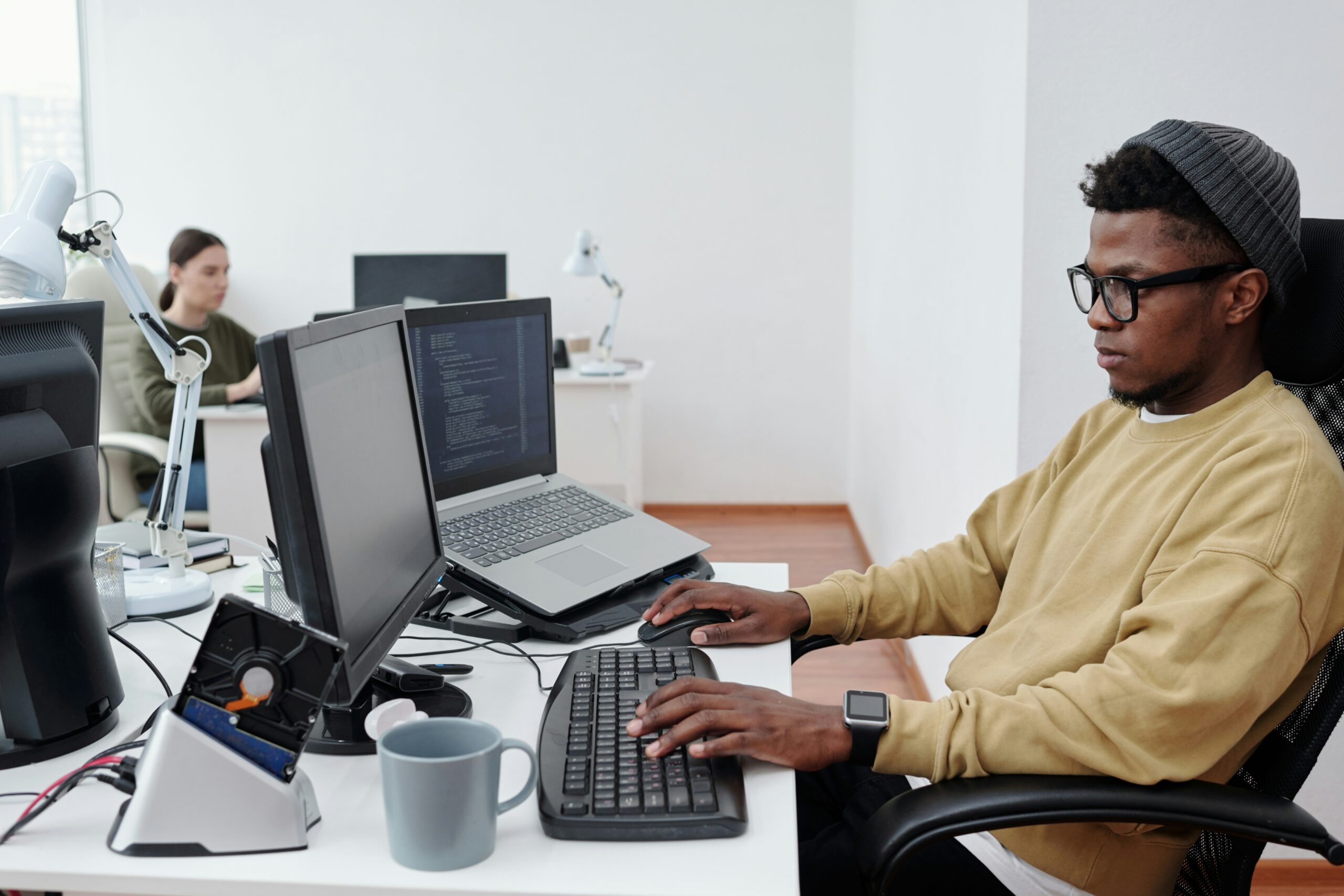 A young man in glasses and a beanie works on IT consulting at a computer in an office.