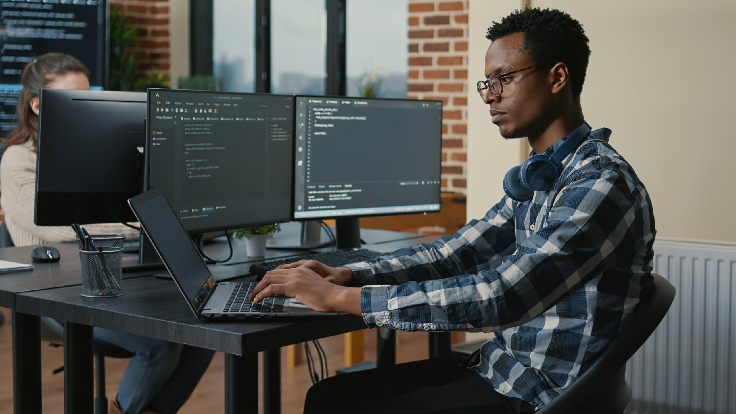 A young man with headphones around his neck works on a laptop in an office with dual monitors, coding to provide a software as part of MSP service.