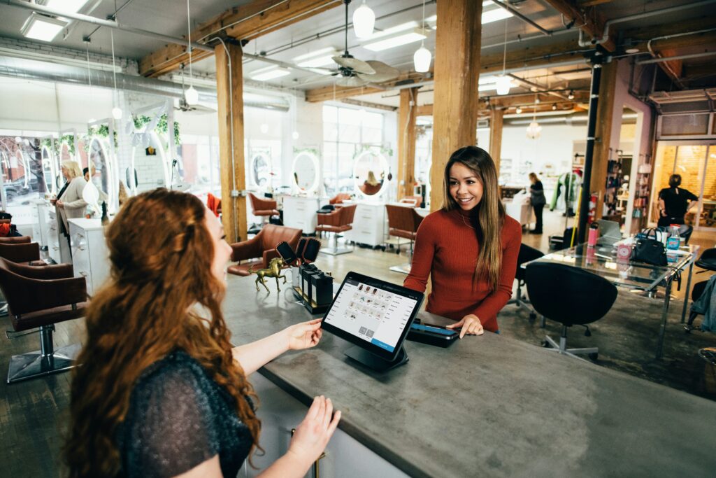 A smiling receptionist assists a customer at the checkout counter in a modern hair salon, proudly supported by their managed IT services for small businesses.