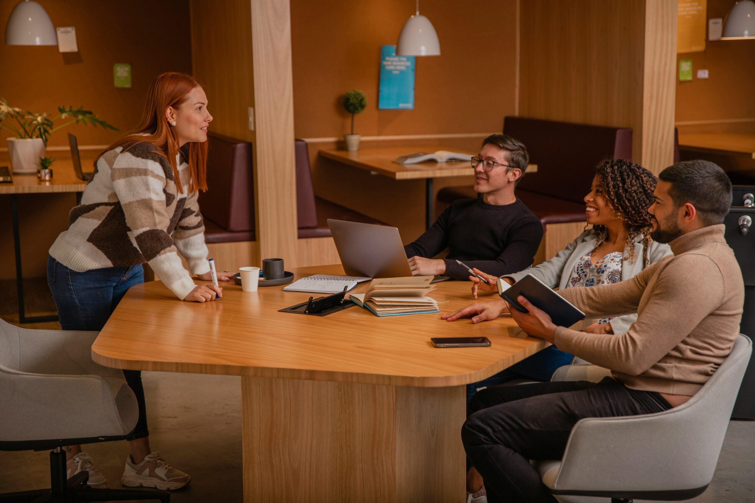 Four adults engaging in a meeting around a wooden table in a warmly lit office space, using laptops and notebooks to discuss what makes a good IT consultant.