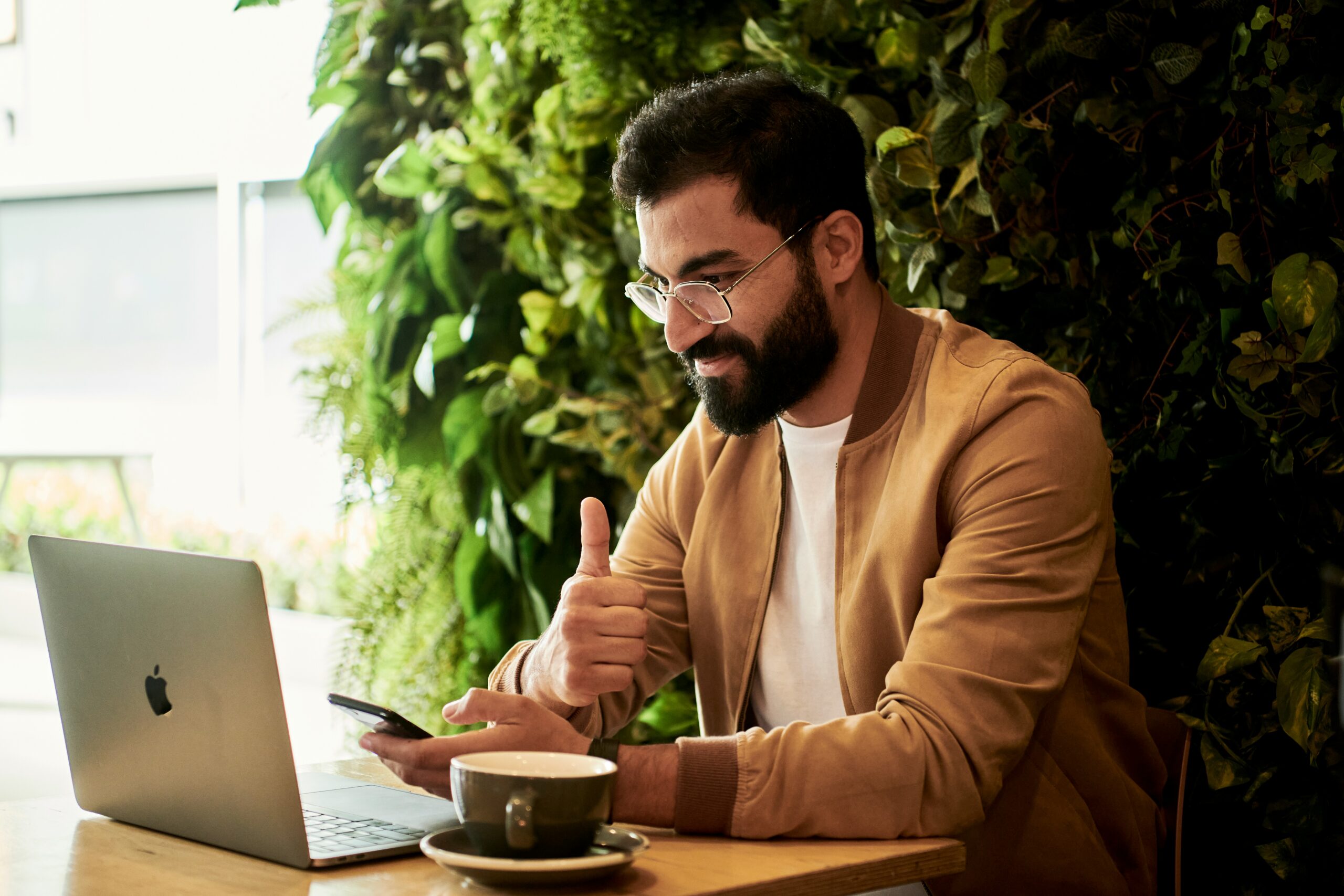 Man in glasses and a tan jacket using a laptop and smartphone at a cafe table, giving a thumbs up after virtual meeting on what is fully managed IT services.