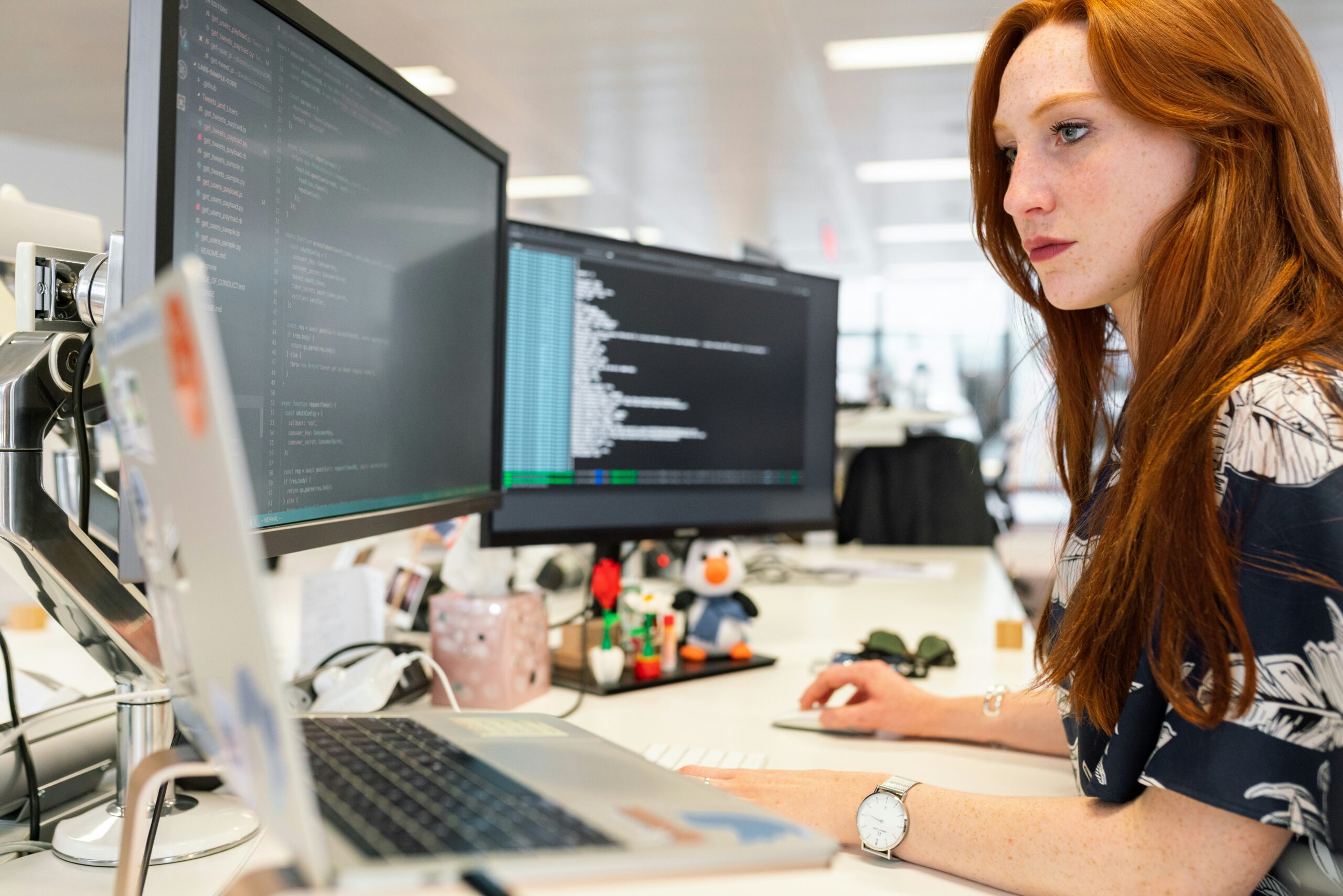 Red-haired woman works at a desk with multiple computer screens displaying code in a modern office specializing in fully managed IT services.