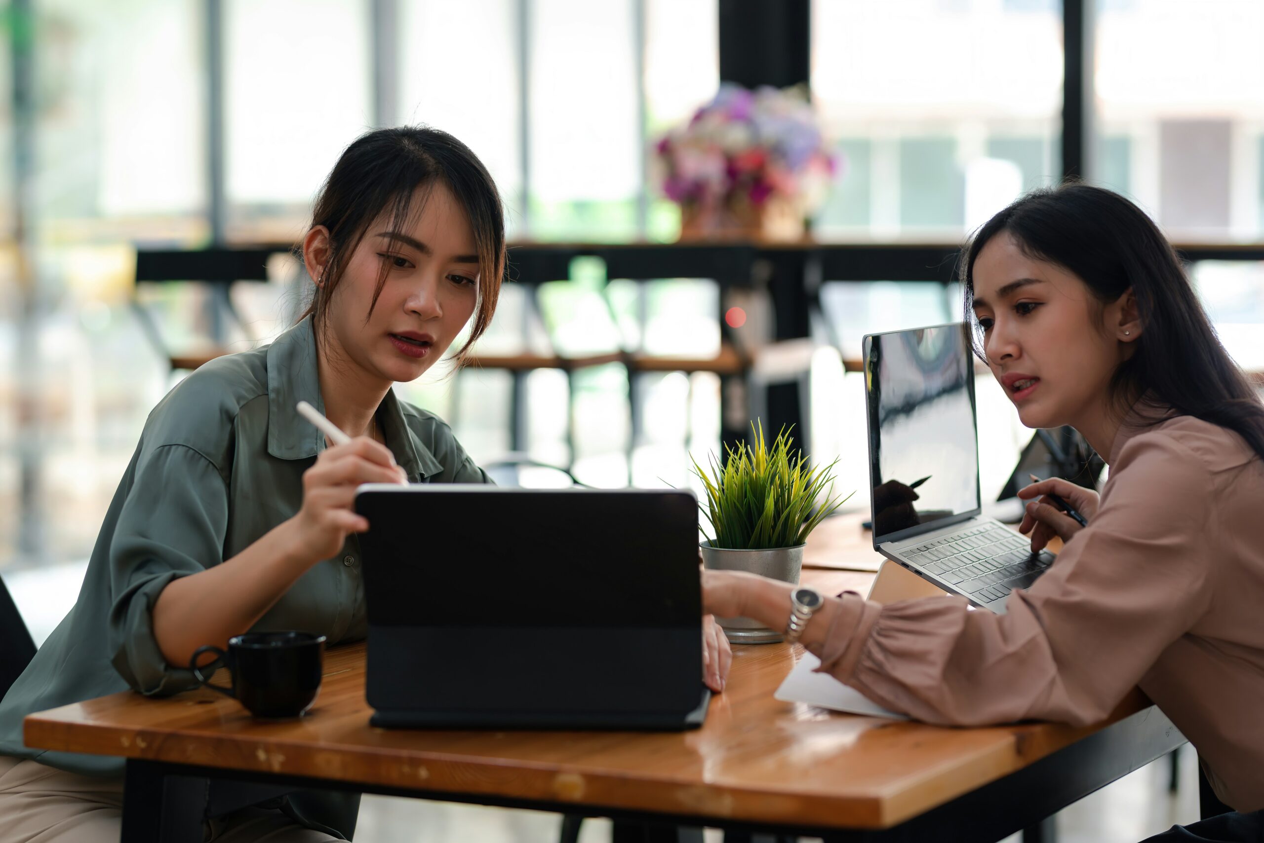 Two women, one an IT consultant, working on laptops in a modern office setting, discussing over digital tablets with coffee and indoor plants on the table.