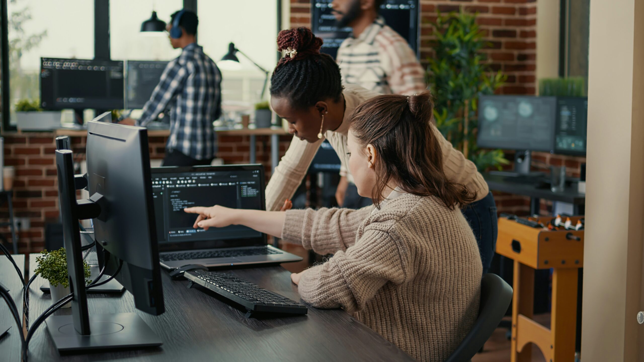 Two women working on a computer in a busy MSP service environment, discussing code on the screen, with other colleagues in the background.