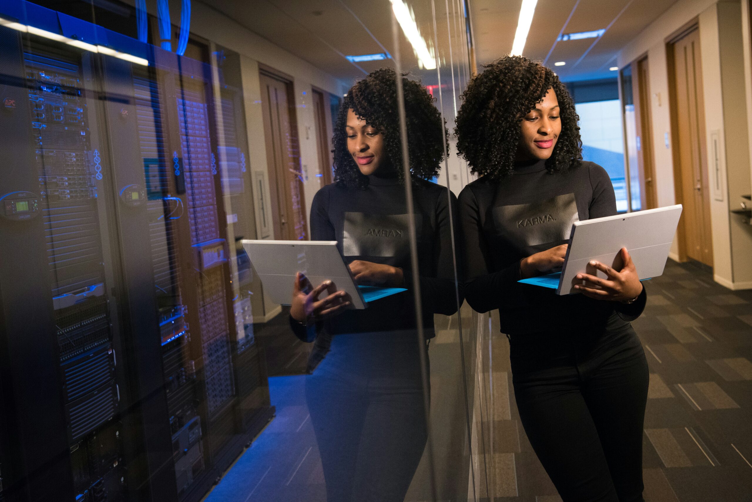 A woman in a black turtleneck checks a tablet for insights on why tech consulting is essential while standing beside a glass wall reflecting her image, in a server room.