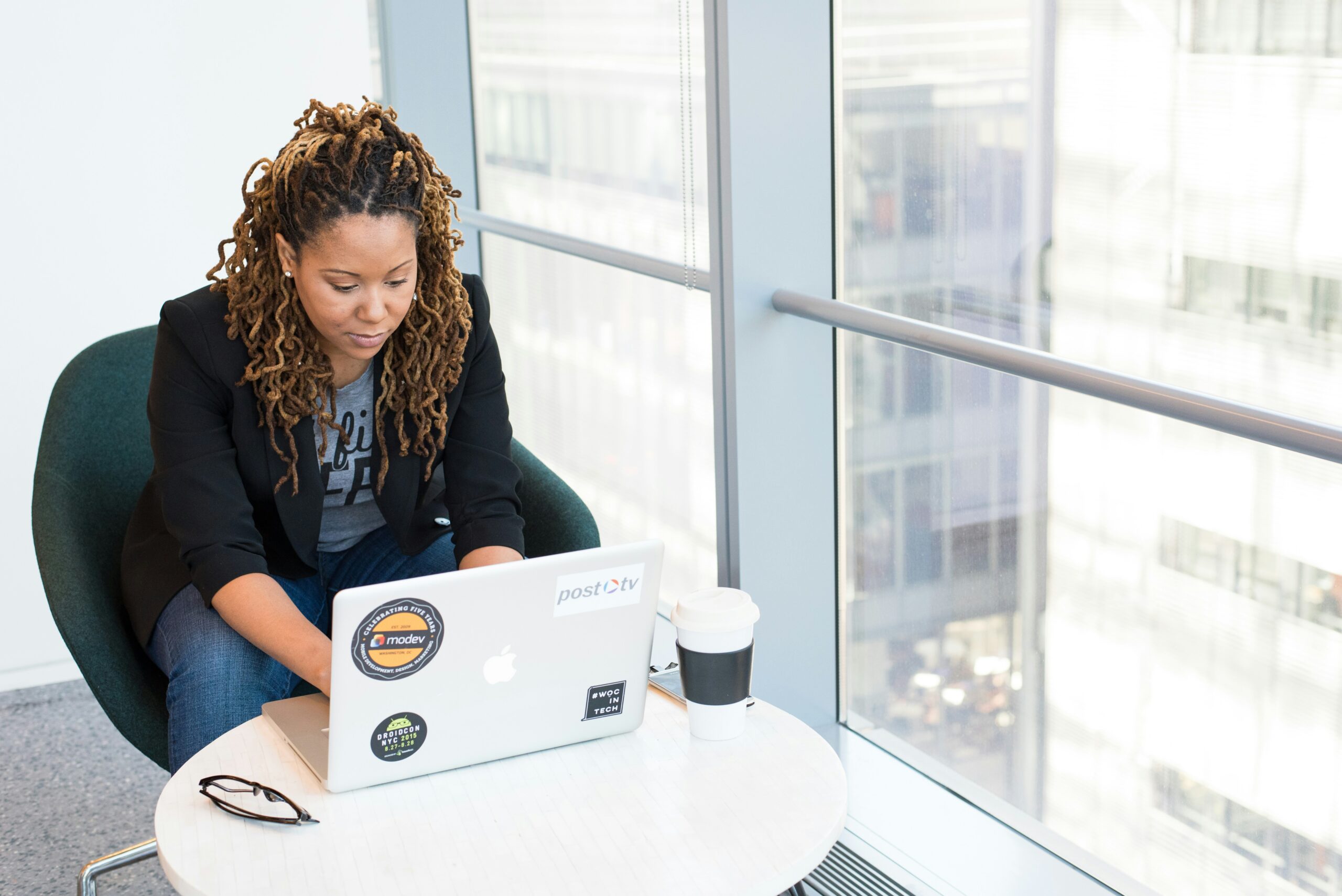 A woman sits at a small table in an office, working on a laptop adorned with stickers, with a coffee cup beside her and a city view through the window, focusing on why tech consulting is pivotal.