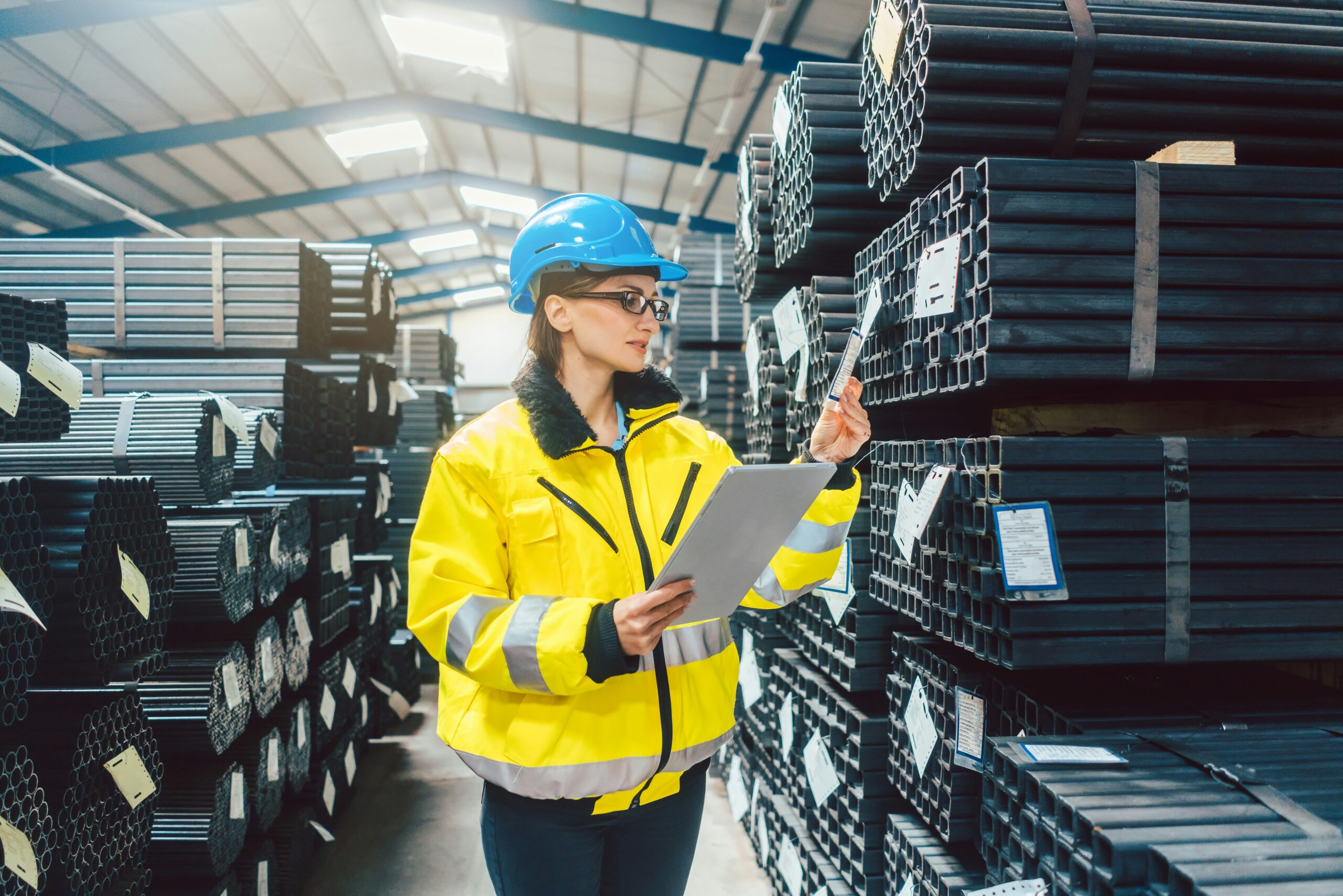 A worker in a high-visibility jacket and blue helmet holds a clipboard and inspects metal pipes stacked in rows inside a warehouse, showcasing the need for quality checks even as industries explore what are examples of process automation.