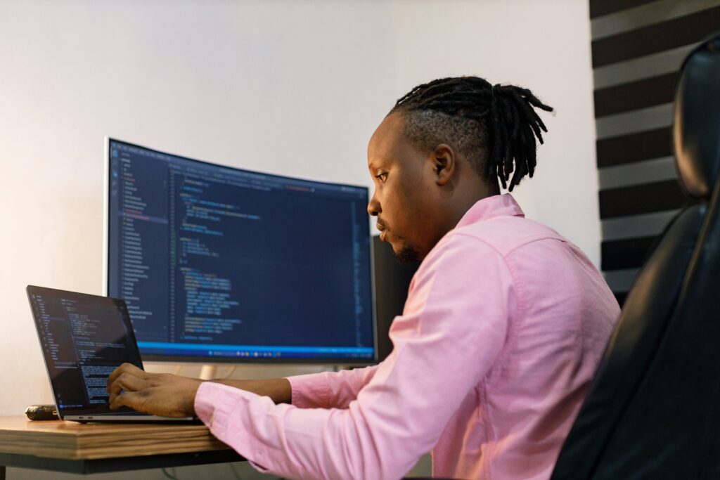 A man in a pink shirt is typing on a laptop, with a large curved monitor displaying coding text related to business process automation software in the background.