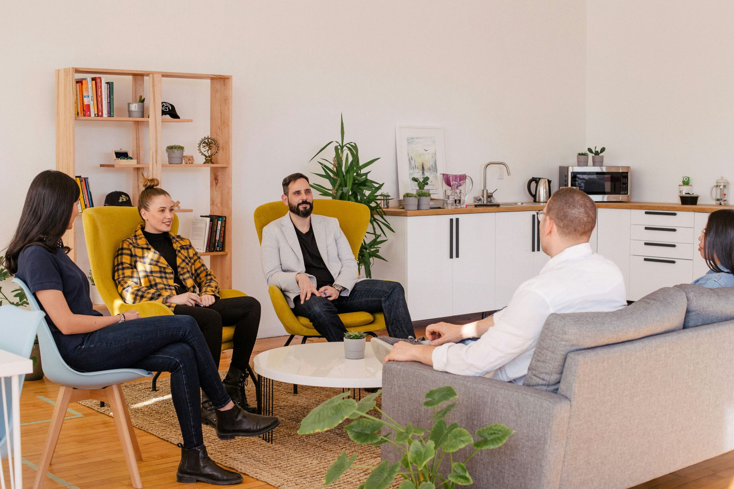 Five adults engage in a conversation about why tech consulting is crucial in a modern office lounge with chairs, a sofa, and plants, creating a relaxed, collaborative atmosphere.