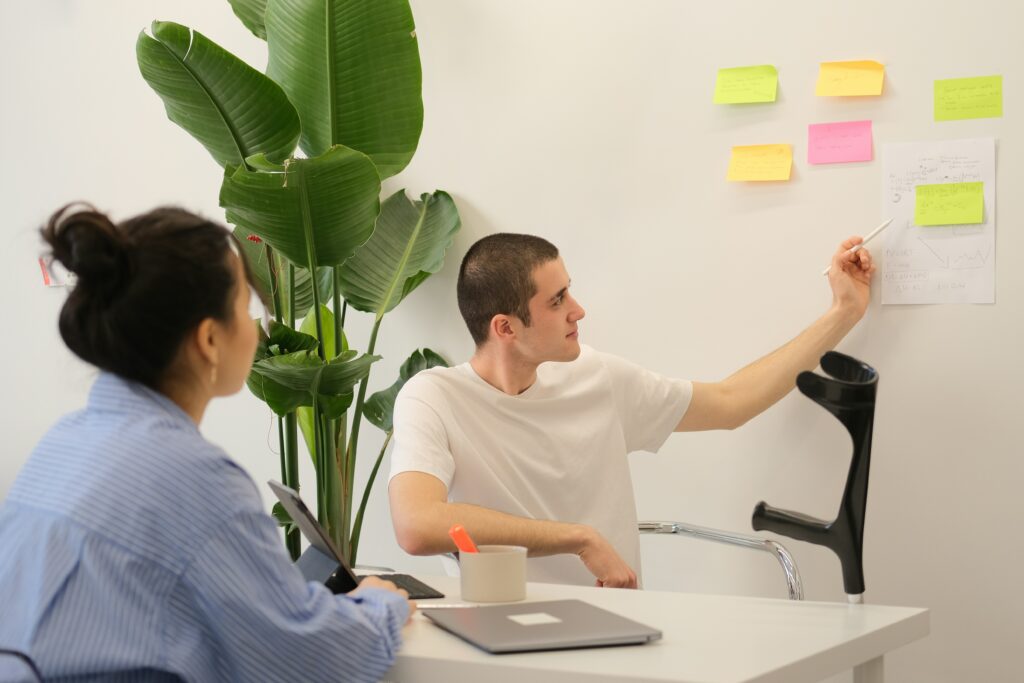 Two people are in an office with one presenting ideas on sticky notes and papers on the wall, integrating concepts of business process automation software, while the other listens attentively.