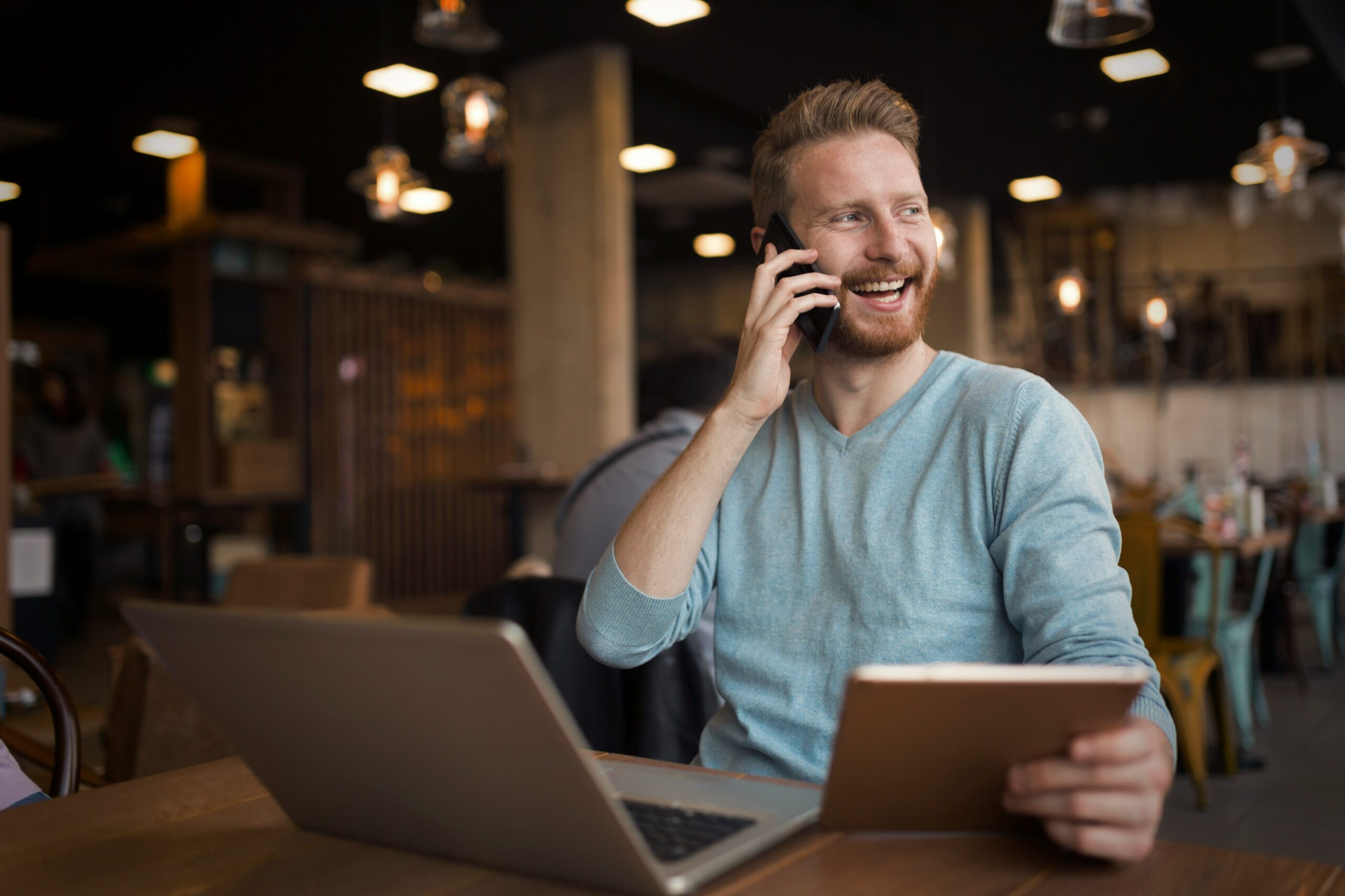 Man smiling while talking on the phone, sitting in a cafe with a laptop and tablet, discussing his example of BPA in his business.