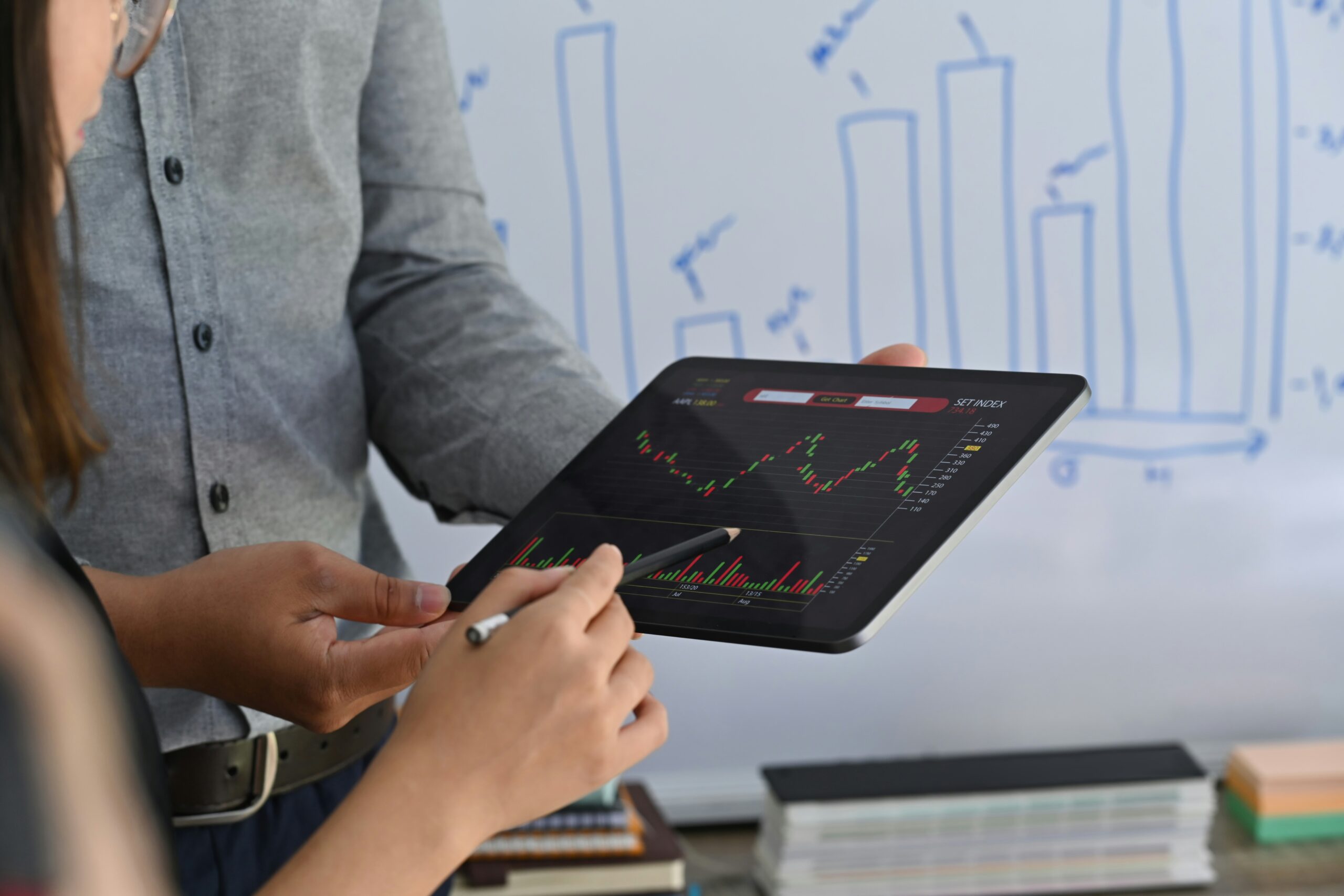 Two individuals analyze stock market data displayed on a tablet in front of a whiteboard with bar charts, discussing how process automation would have made analysis easier.