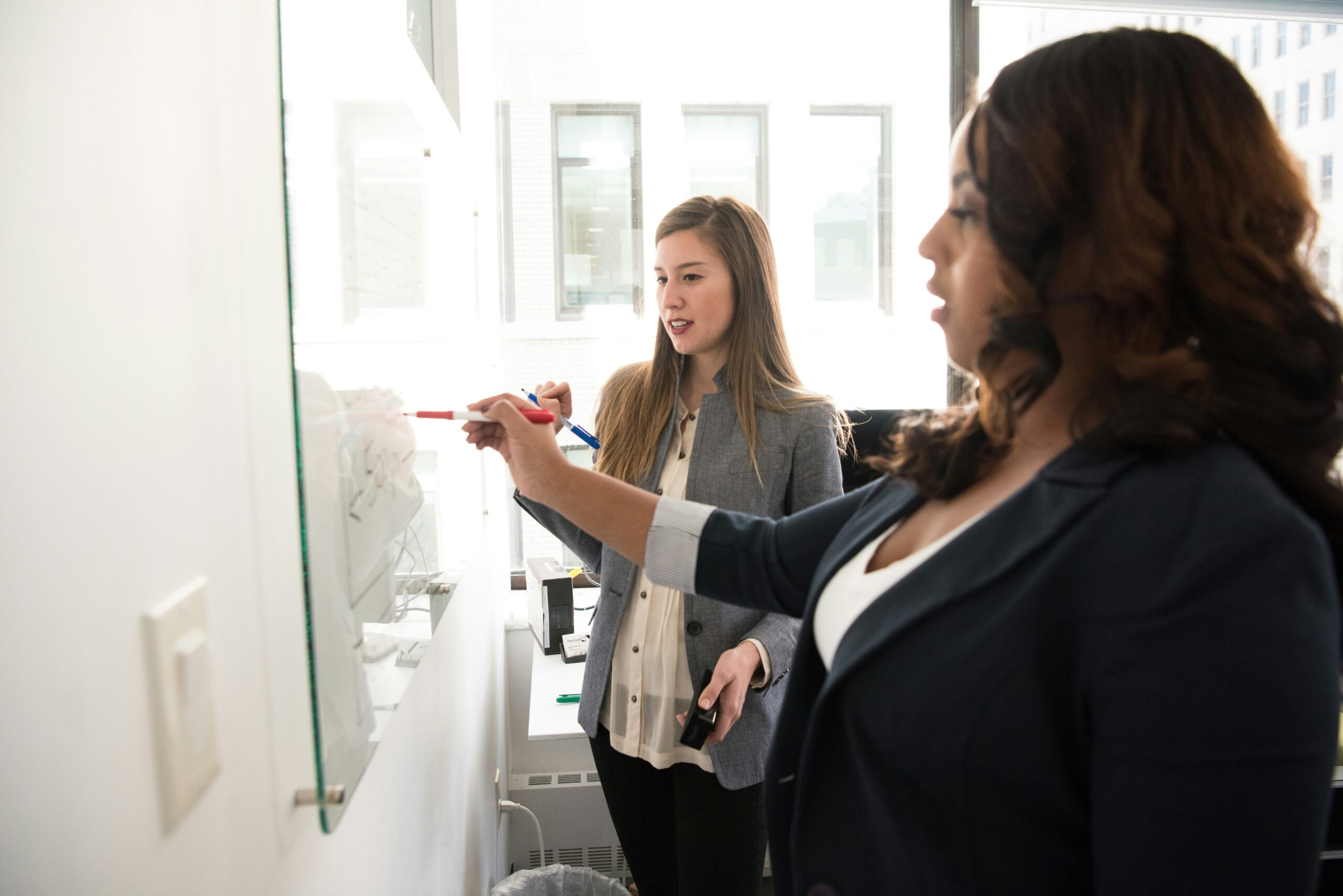 Two professional women collaborate at a whiteboard in a tech consulting office, with one writing and explaining as the other listens attentively.