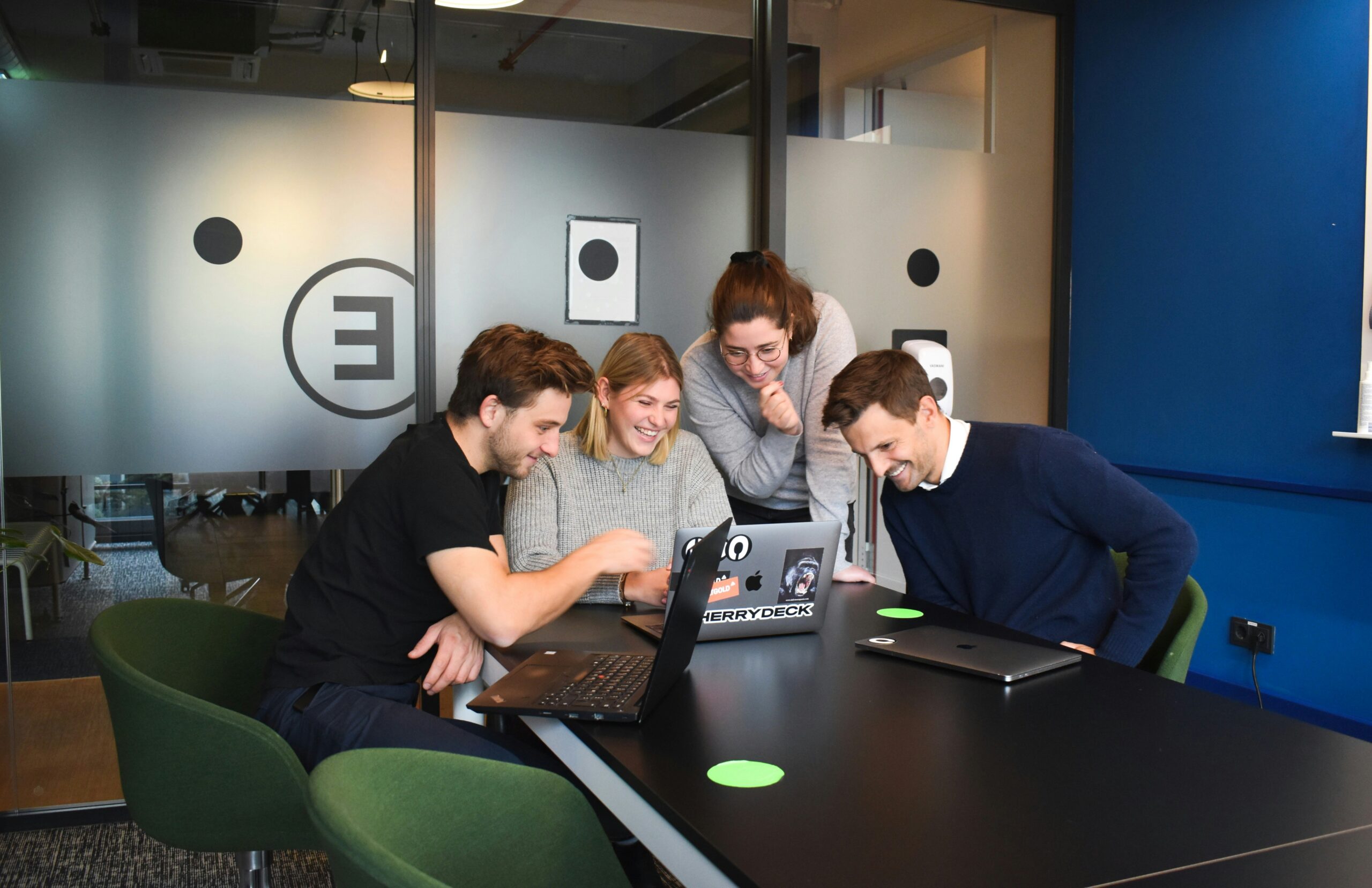 A group of five people are gathered around a laptop in an office setting, smiling and looking at the screen, perhaps discovering how to automate your business processes.