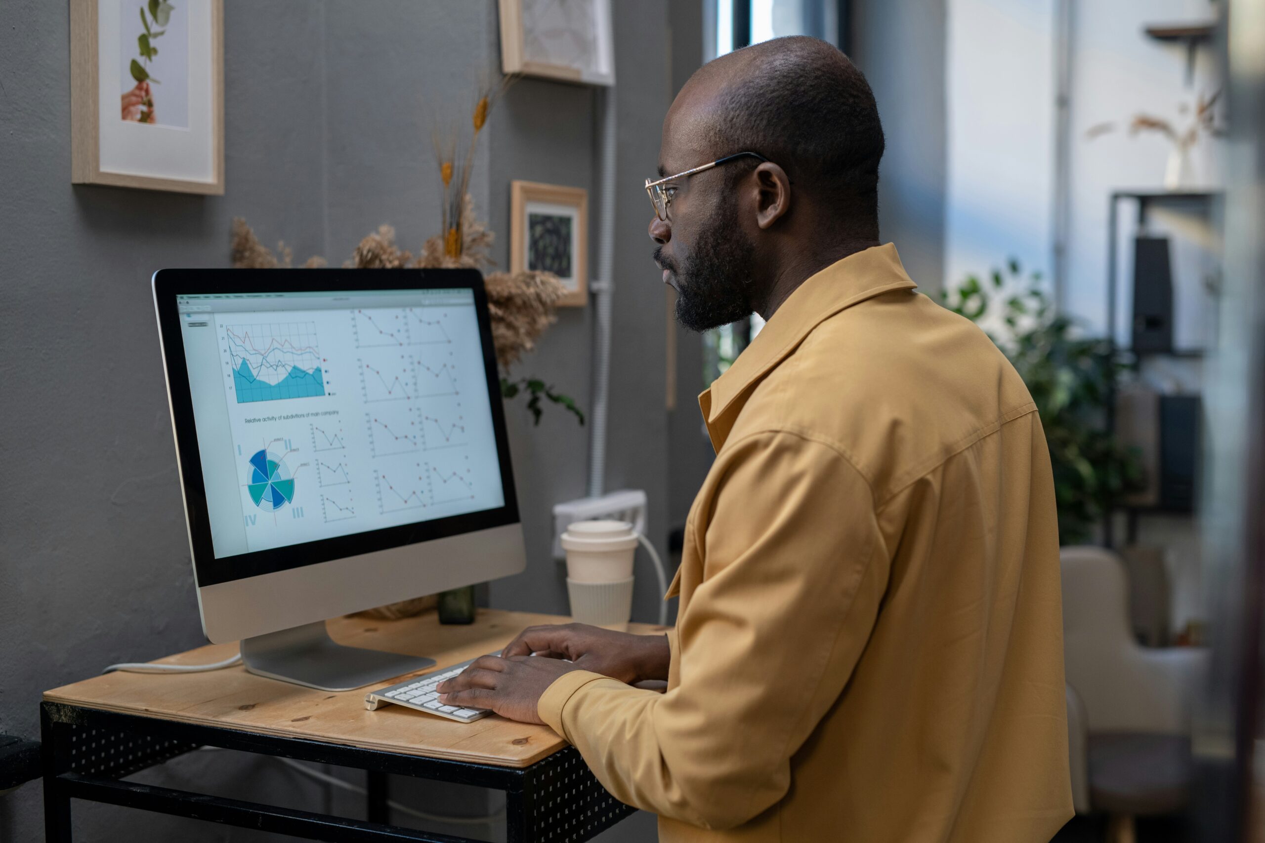A man wearing glasses and a yellow shirt analyzes graphs and charts on a computer screen at a standing desk, contemplating the impact of automating your business processes.