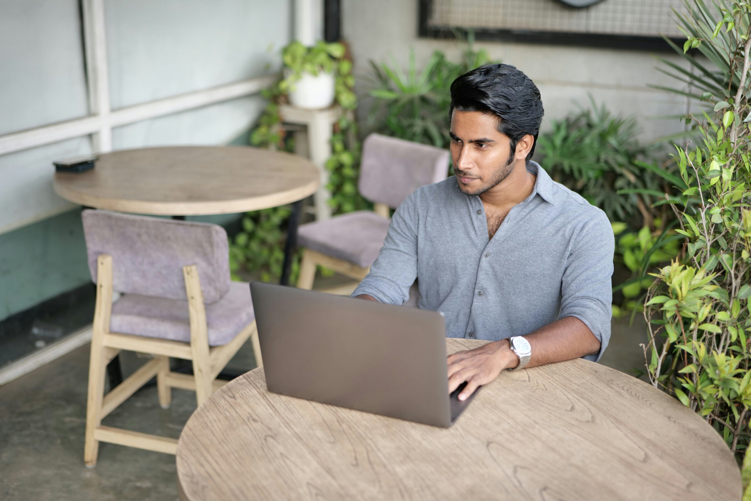 A person is working on a laptop at an outdoor café with wooden tables and green plants in the background, researching how to automate your business processes.