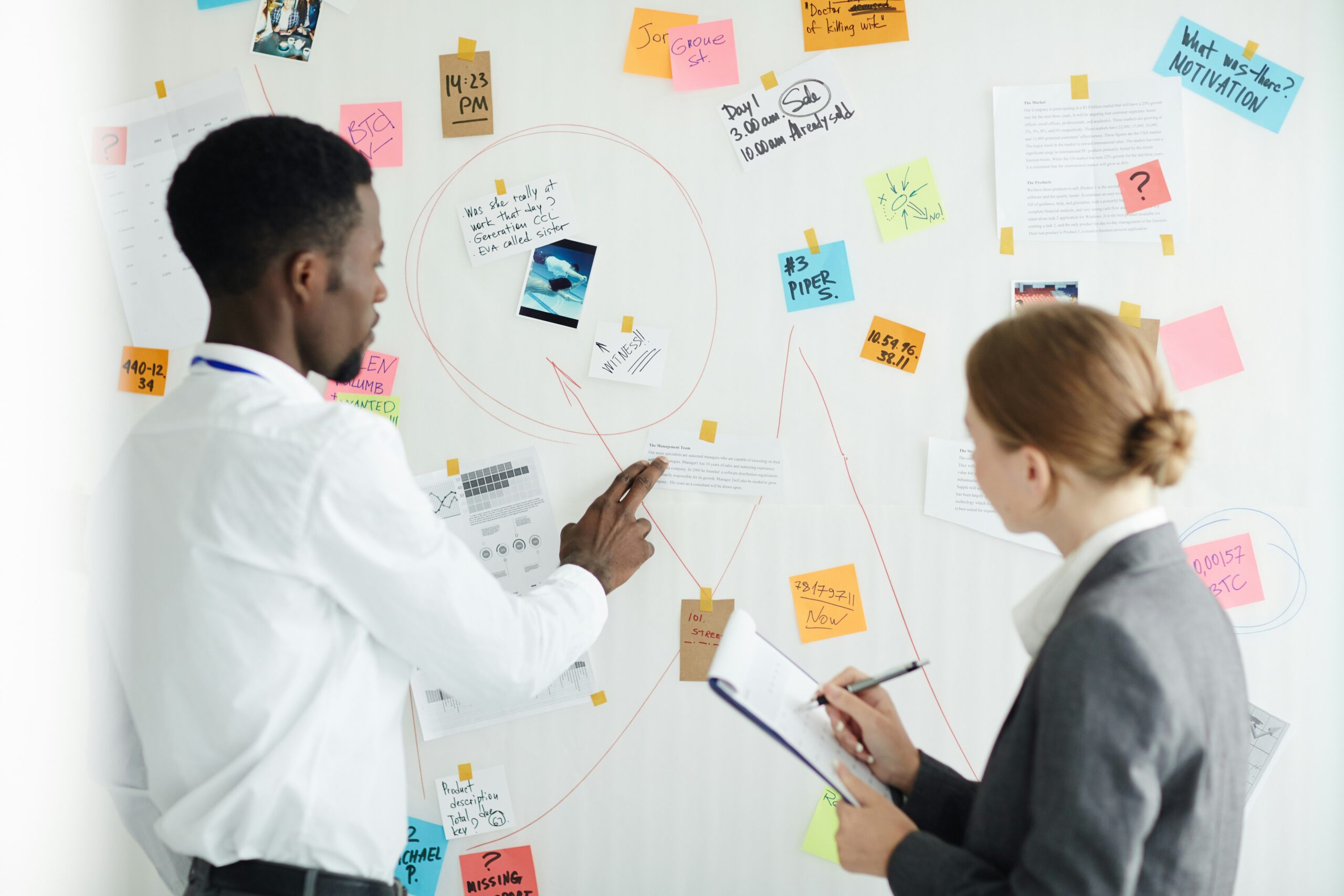 Two people in formal attire discuss data and charts on a whiteboard covered with sticky notes and paper thinking that workflow automation would help them.