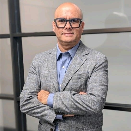A bald man wearing glasses, a grey blazer, and a blue shirt stands with arms crossed in front of a glass wall.
