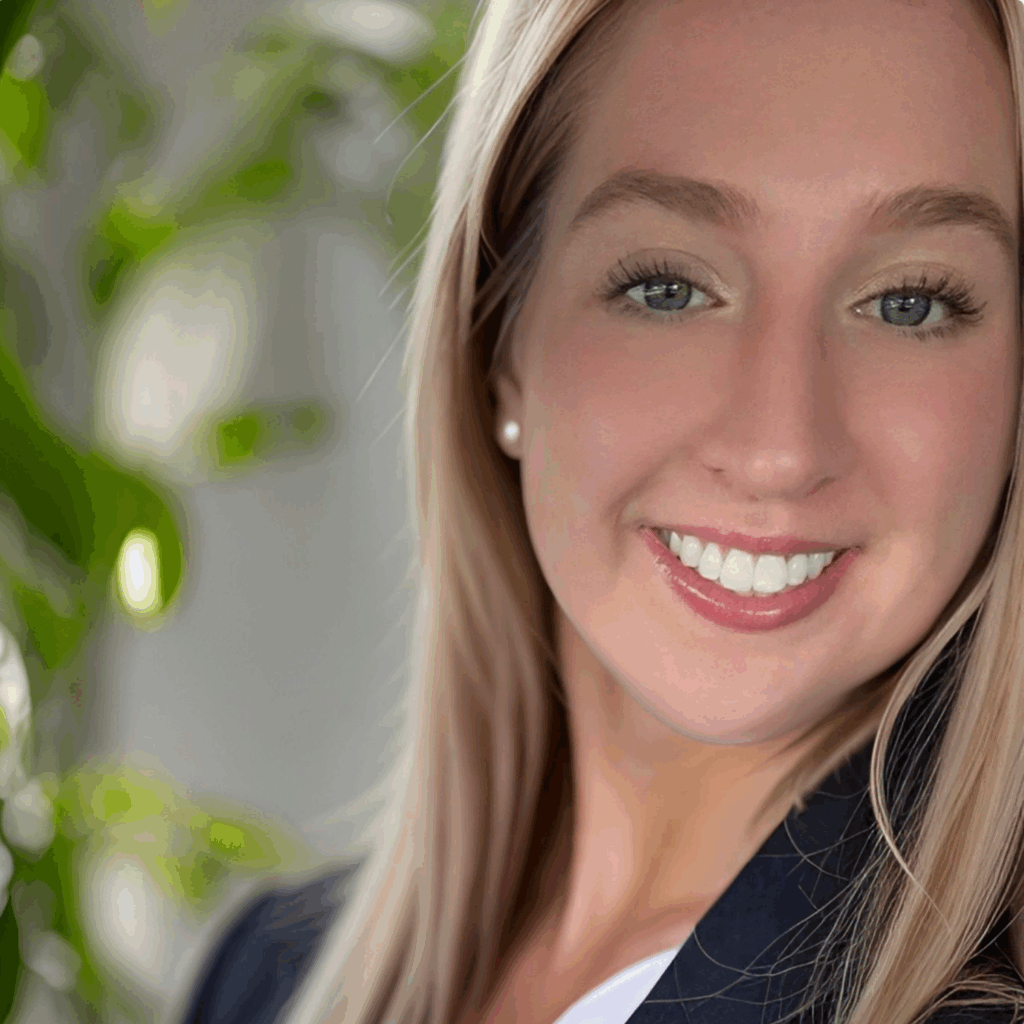 A young woman with long blonde hair smiles at the camera, wearing a black blazer and white top, with a blurred green plant in the background.