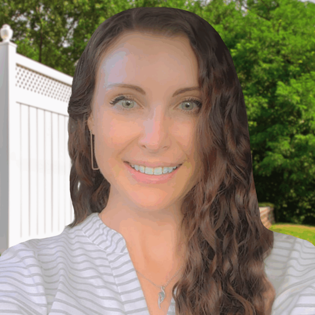 A woman with long wavy brown hair and light eyes smiles outdoors, standing in front of a white fence and green trees.